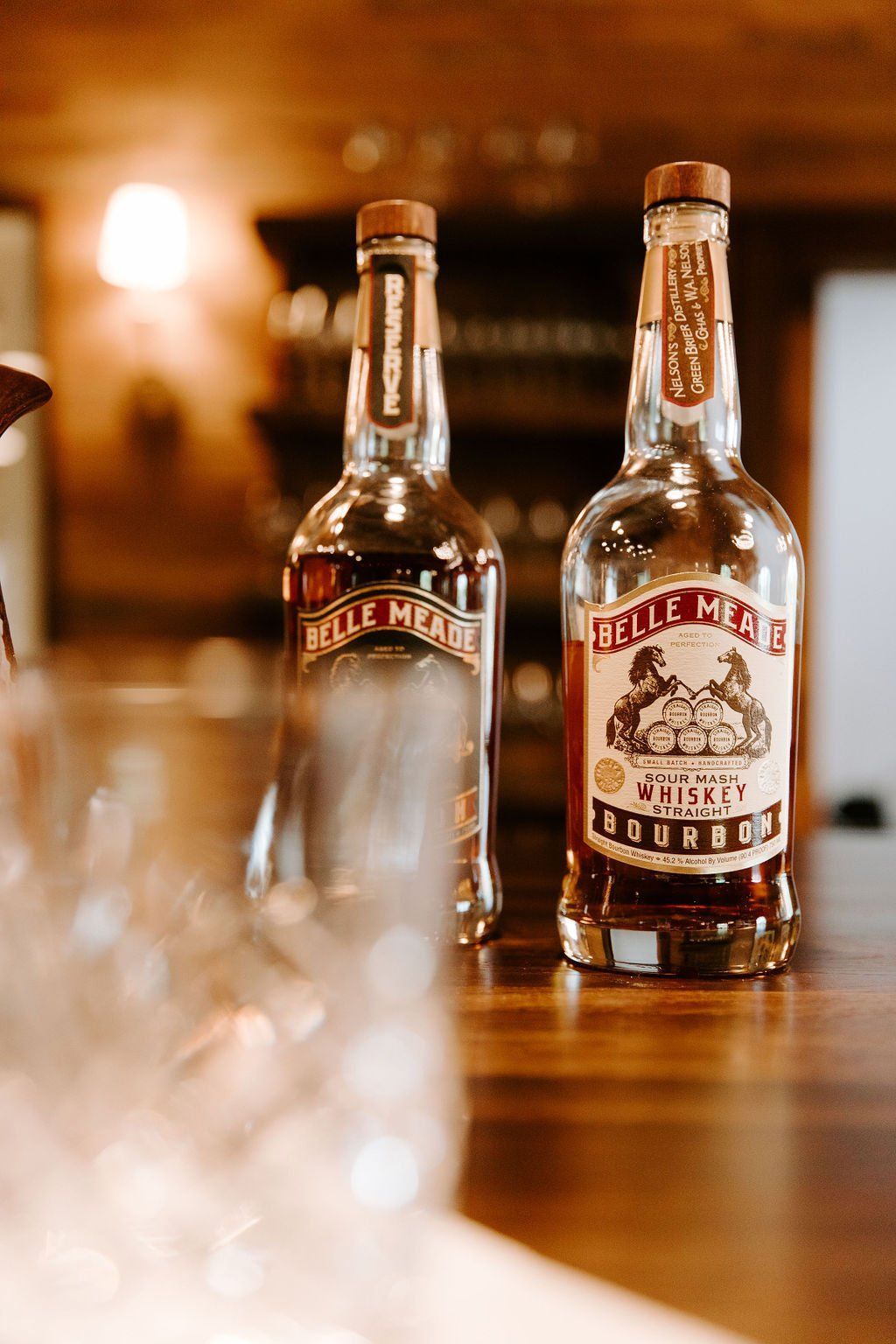Two bottles of Belle Meade Bourbon whiskey on a wooden surface, with blurred glassware in the foreground.