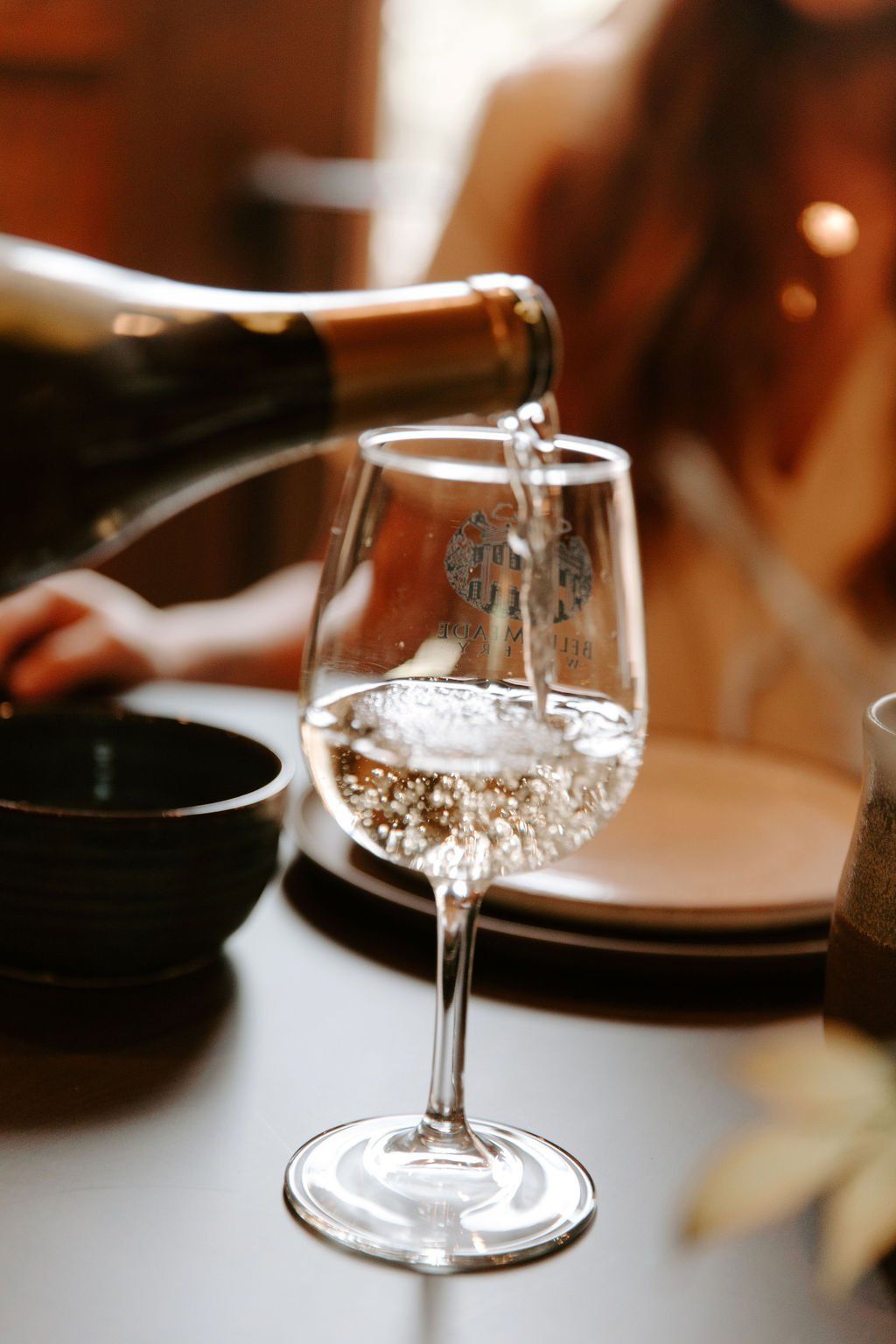 White wine being poured into a clear glass at a table. A person is slightly blurred in the background.