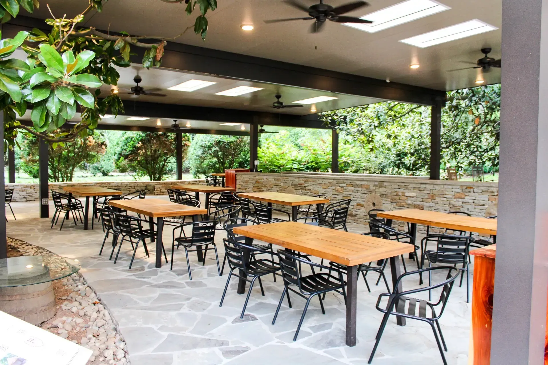 Outdoor dining area with tables and chairs under a covered patio with trees in the background.