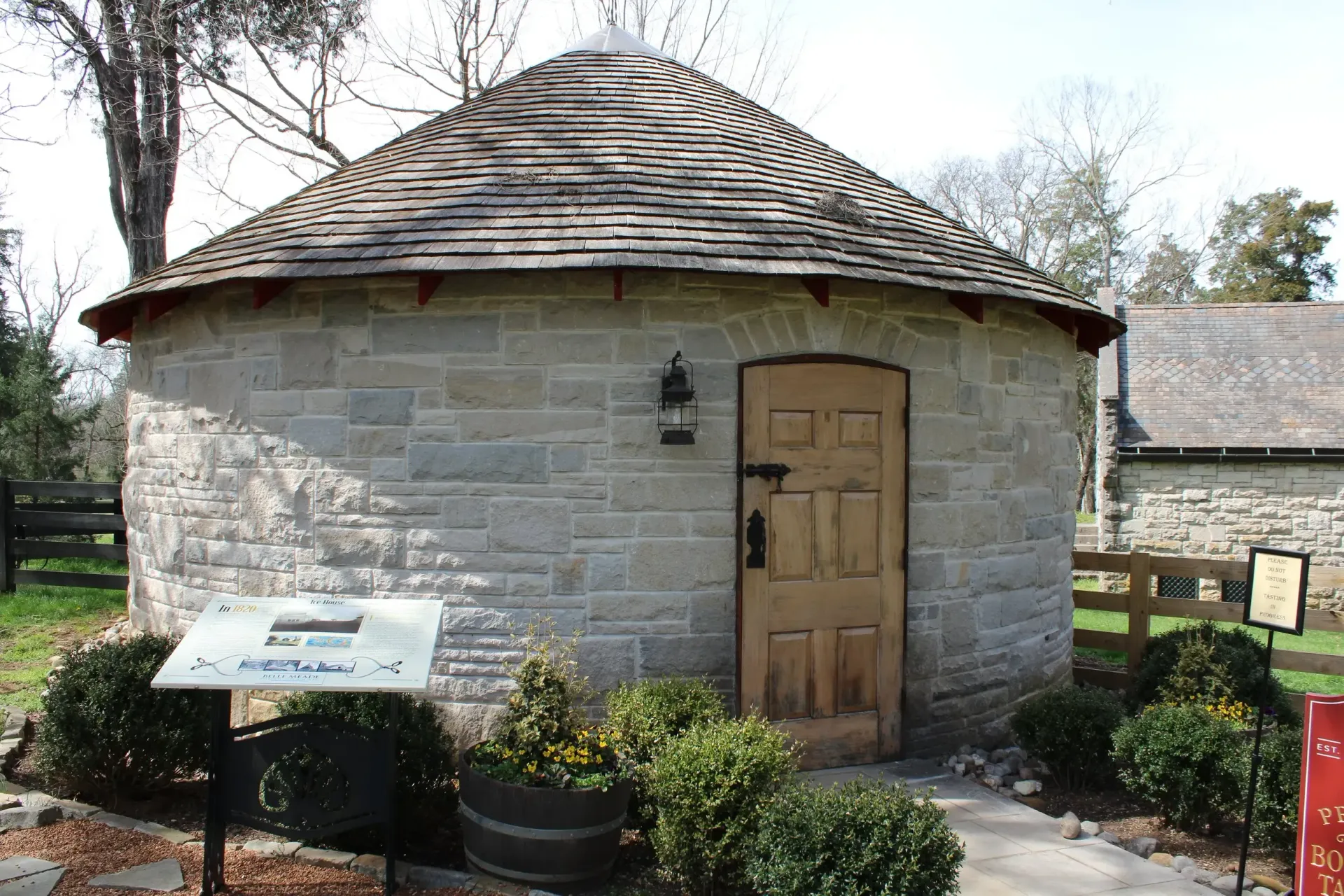 Round stone building with wooden door, tile roof, and a sign in a grassy area.