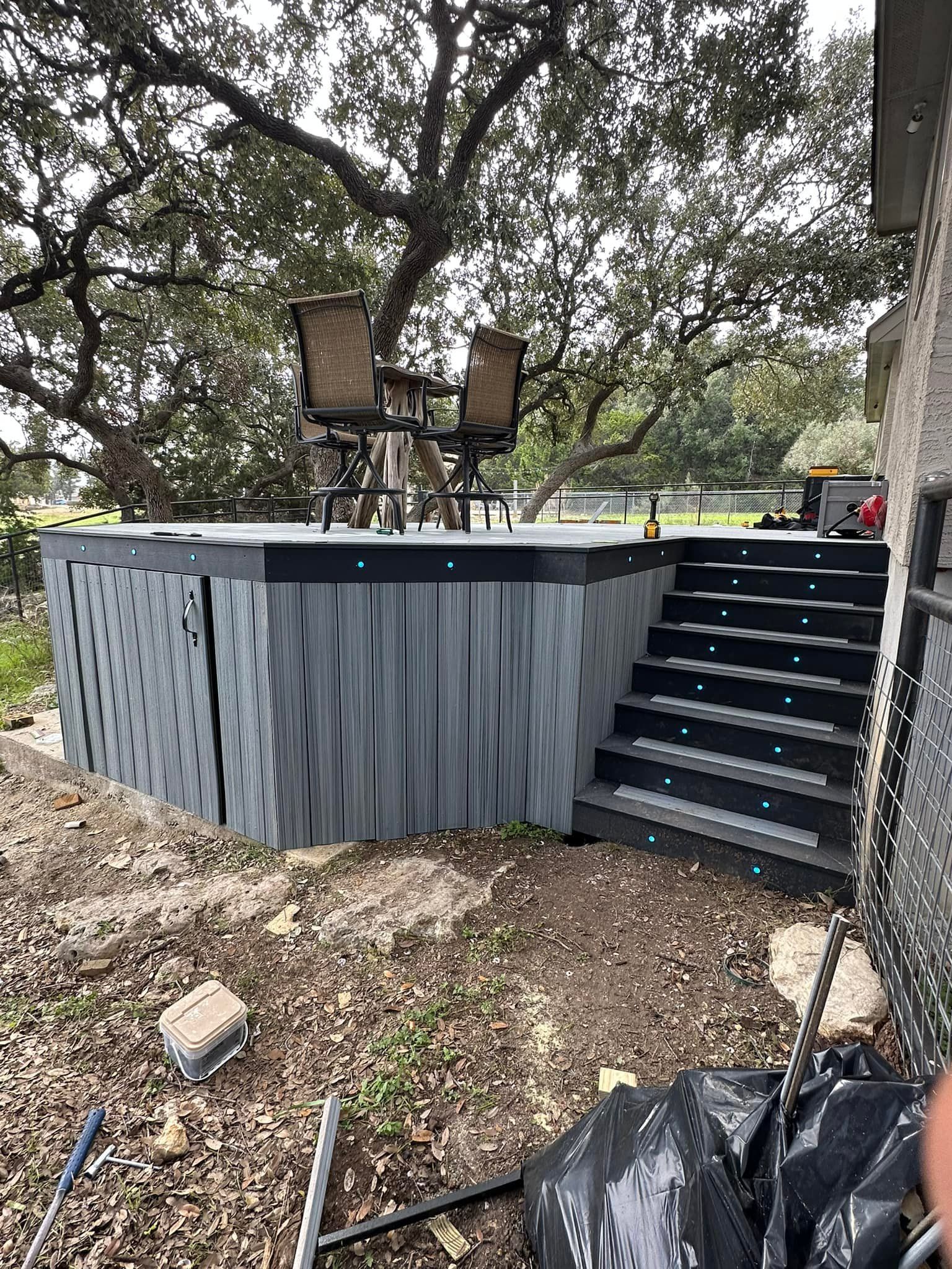 A raised deck with gray siding, black steps, and two bar stools under a tree.