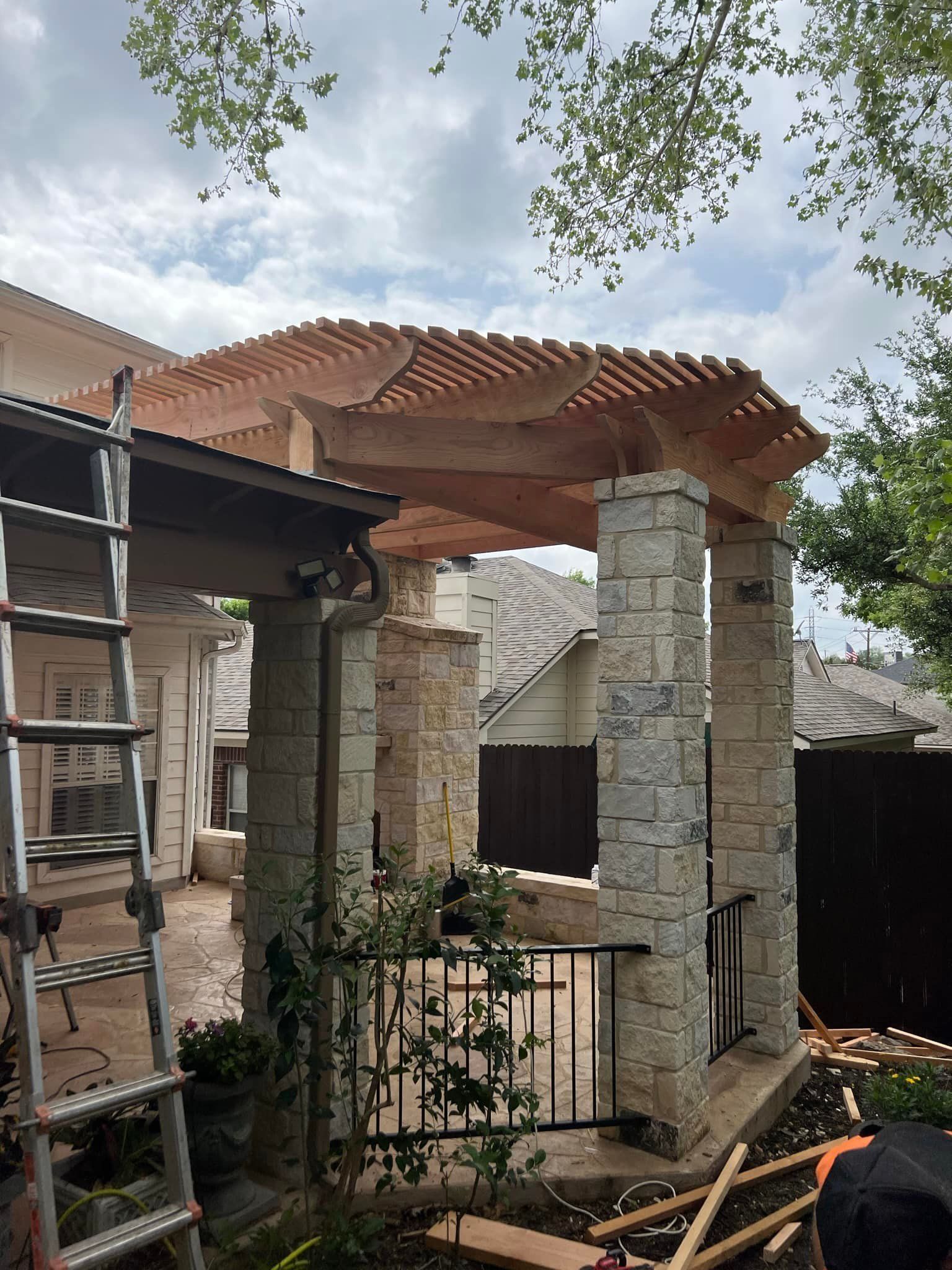 Stone pillar pergola with wooden beams under construction outdoors. A ladder leans against the house.