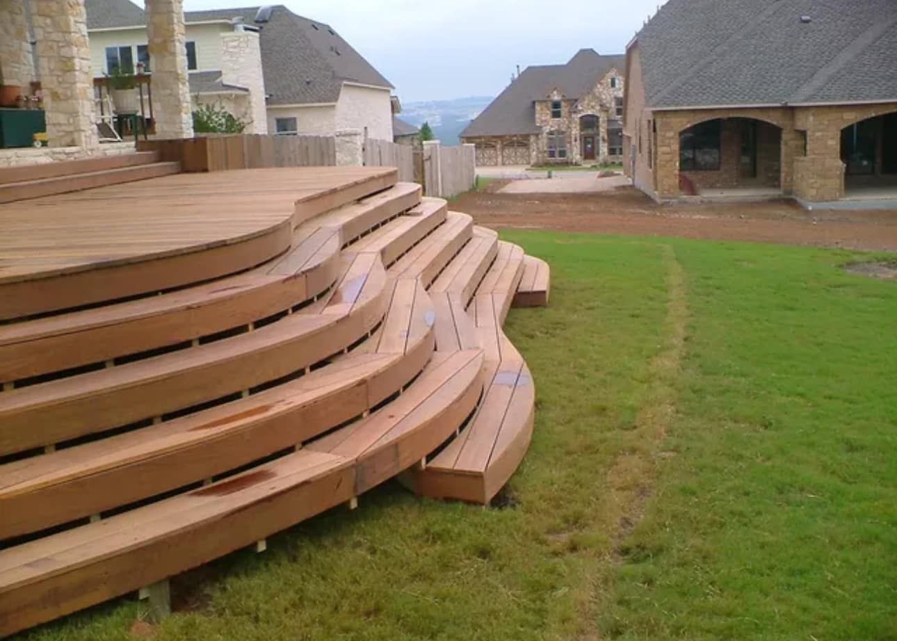 Wooden deck with curved steps leading down to grassy yard, large houses in the background.