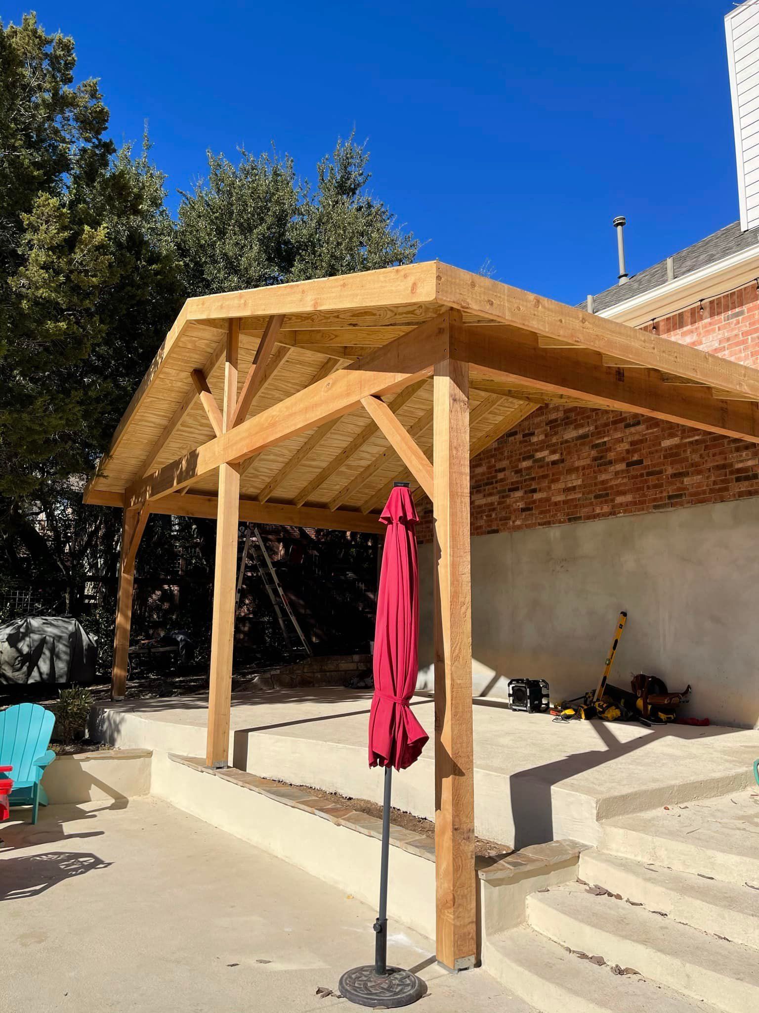 Wooden patio cover over a cement patio with a red umbrella, against a building and trees.