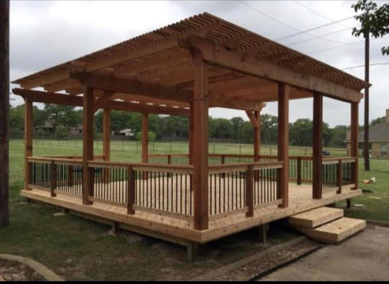 Wooden pergola with deck and railing on a grassy area, overcast sky.