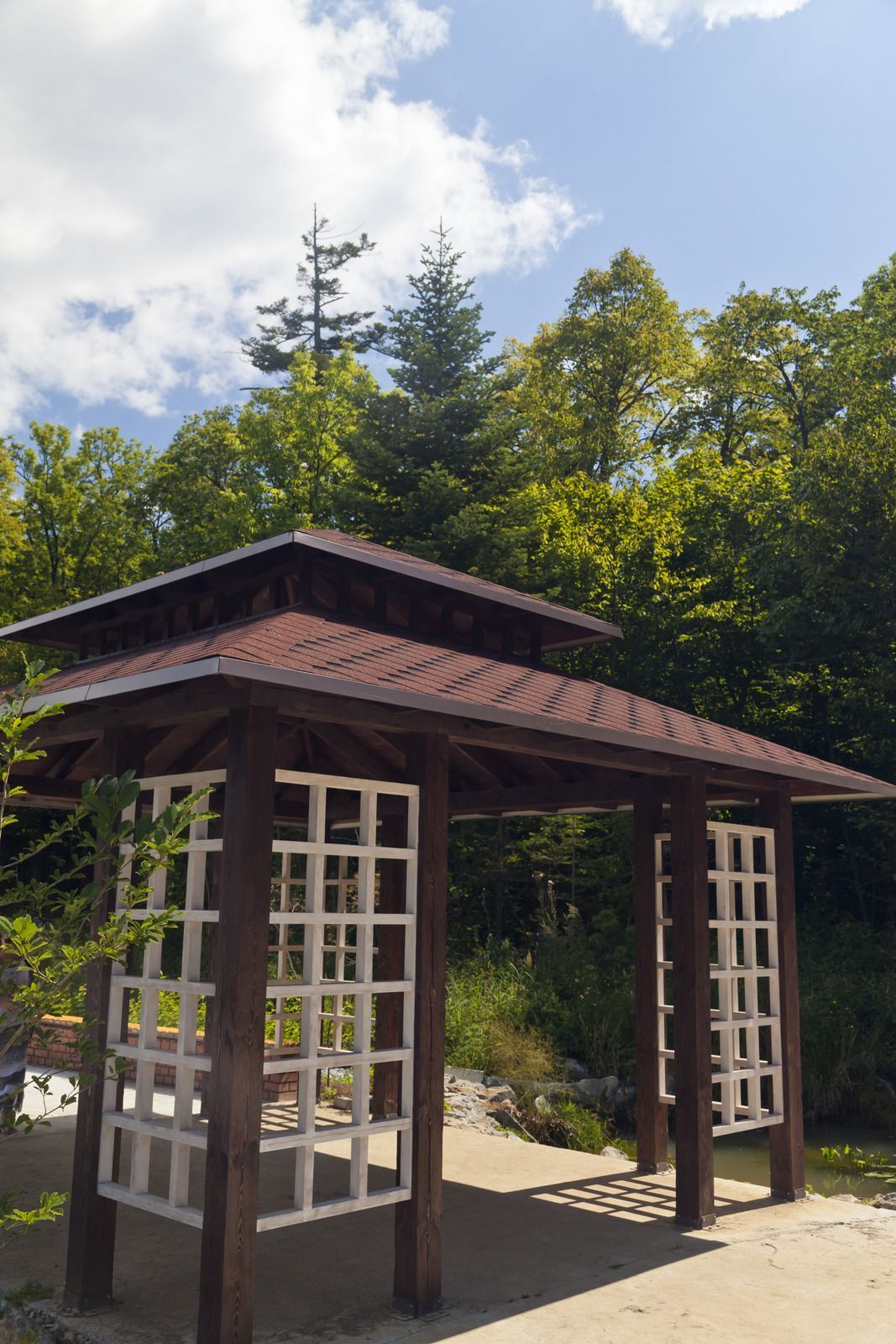 Wooden gazebo with white lattice panels, set in a sunny forest clearing.