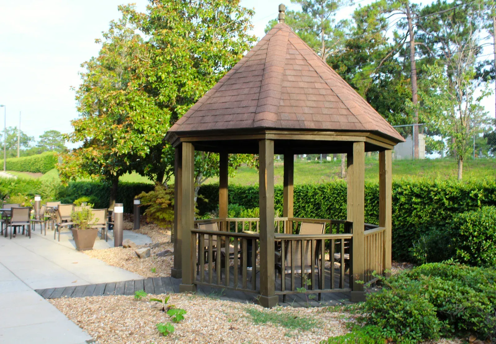 Wooden gazebo with brown roof, surrounded by greenery and seating area.