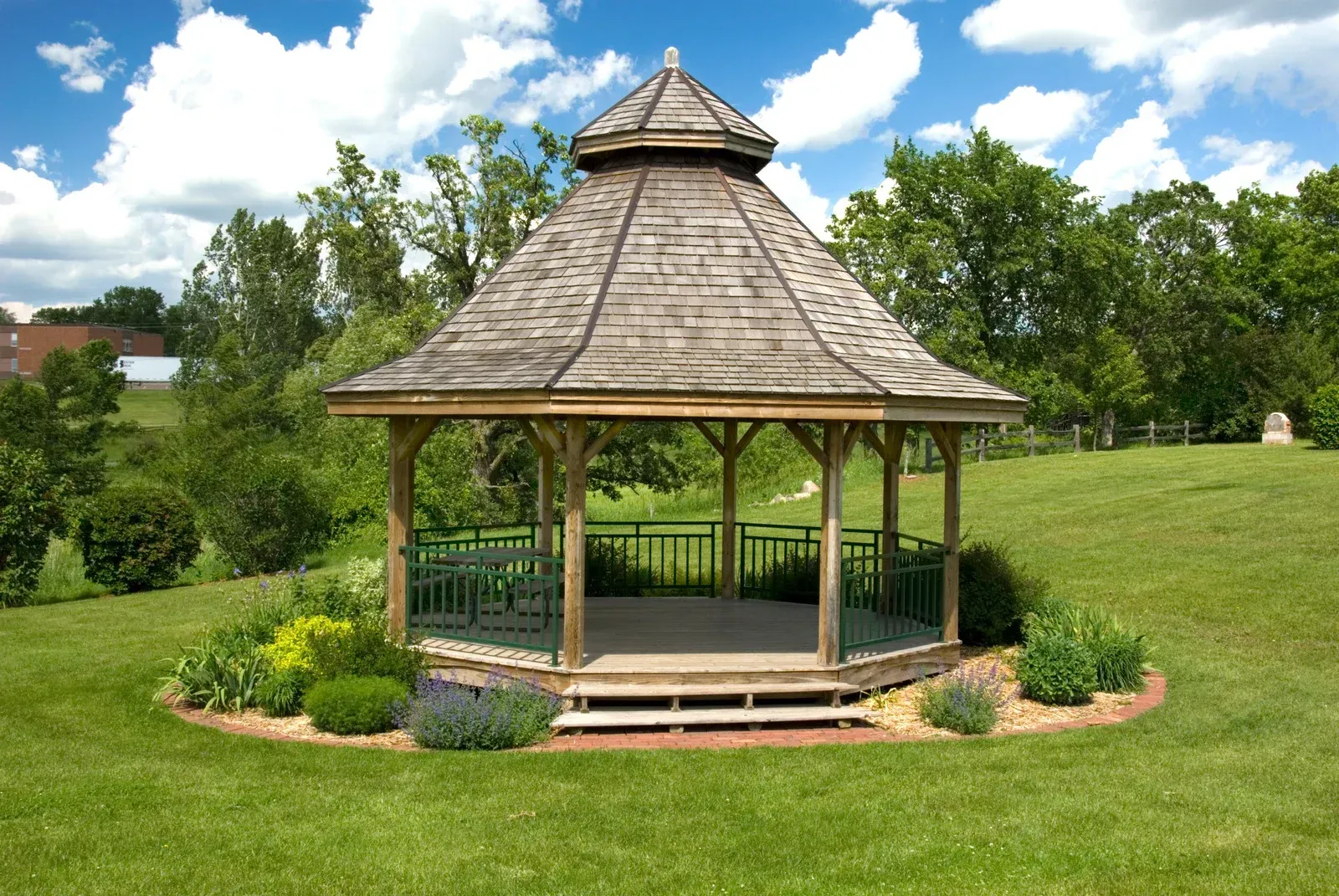 Wooden gazebo with brown roof and green railing in a grassy park, surrounded by plants and trees.