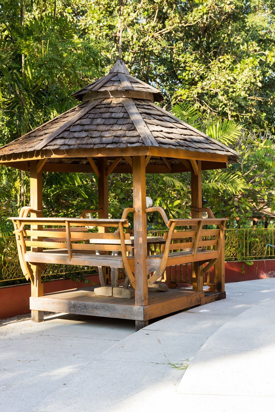 Wooden gazebo with shingle roof, surrounded by trees, on a paved path.