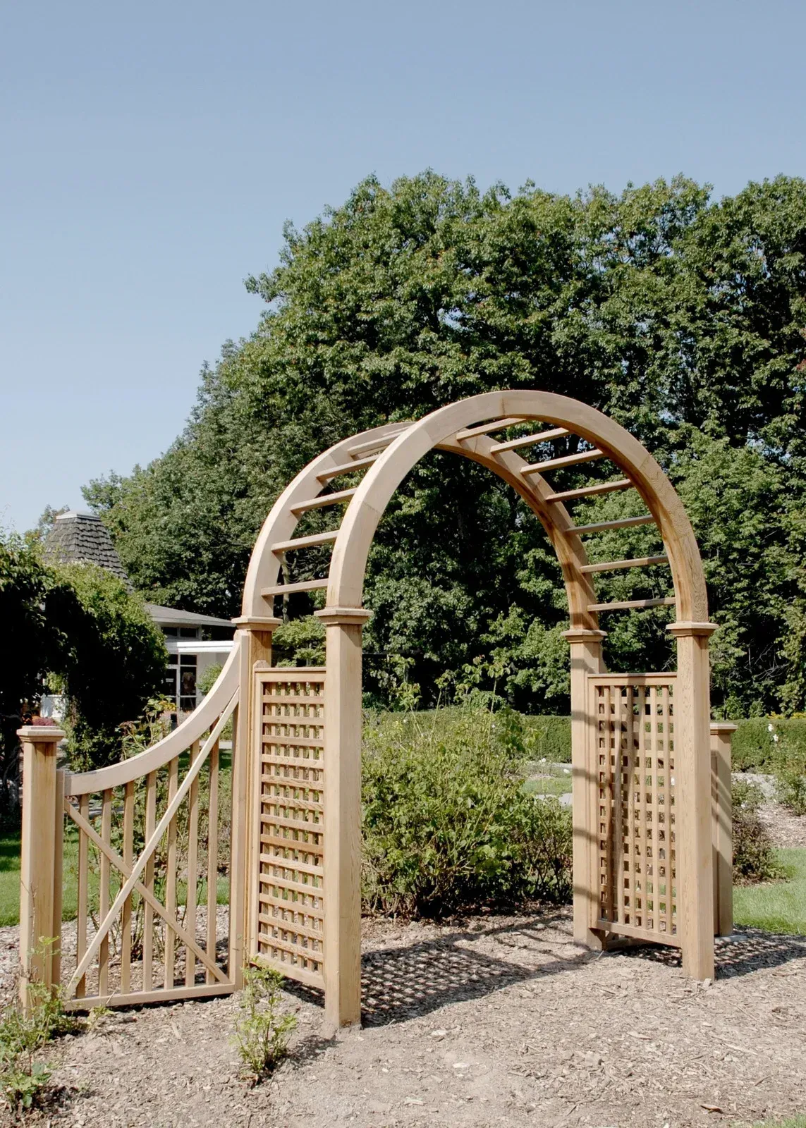 Wooden arched garden gate, surrounded by greenery, under a blue sky.