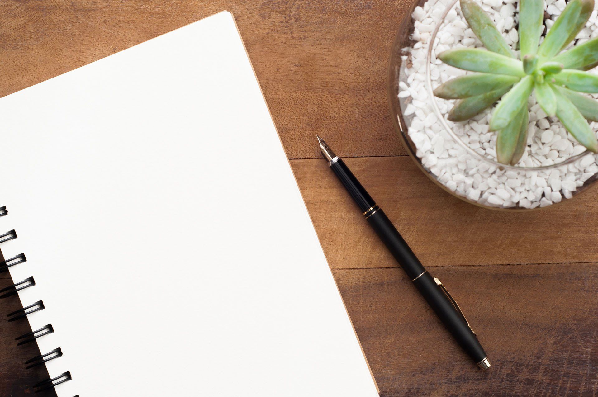 A pen and a notebook are on a wooden table next to a potted plant.