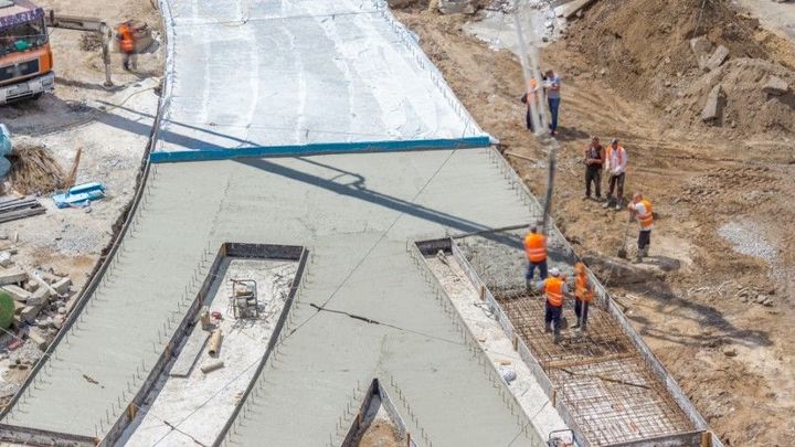 Construction workers pour concrete on a large sloped surface at a roadwork site.