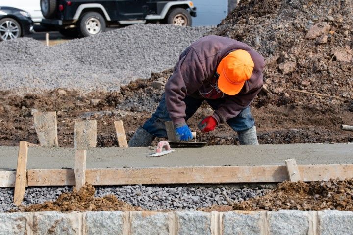 Construction worker smoothing wet concrete at a roadside site, wearing orange hard hat and gloves