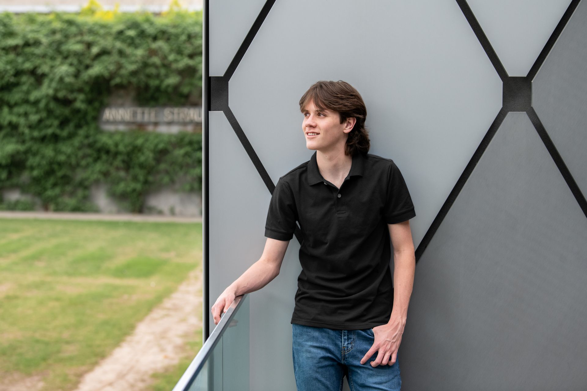 A person in a purple graduation gown and white stole stands outside under a pergola, holding a mortarboard.
