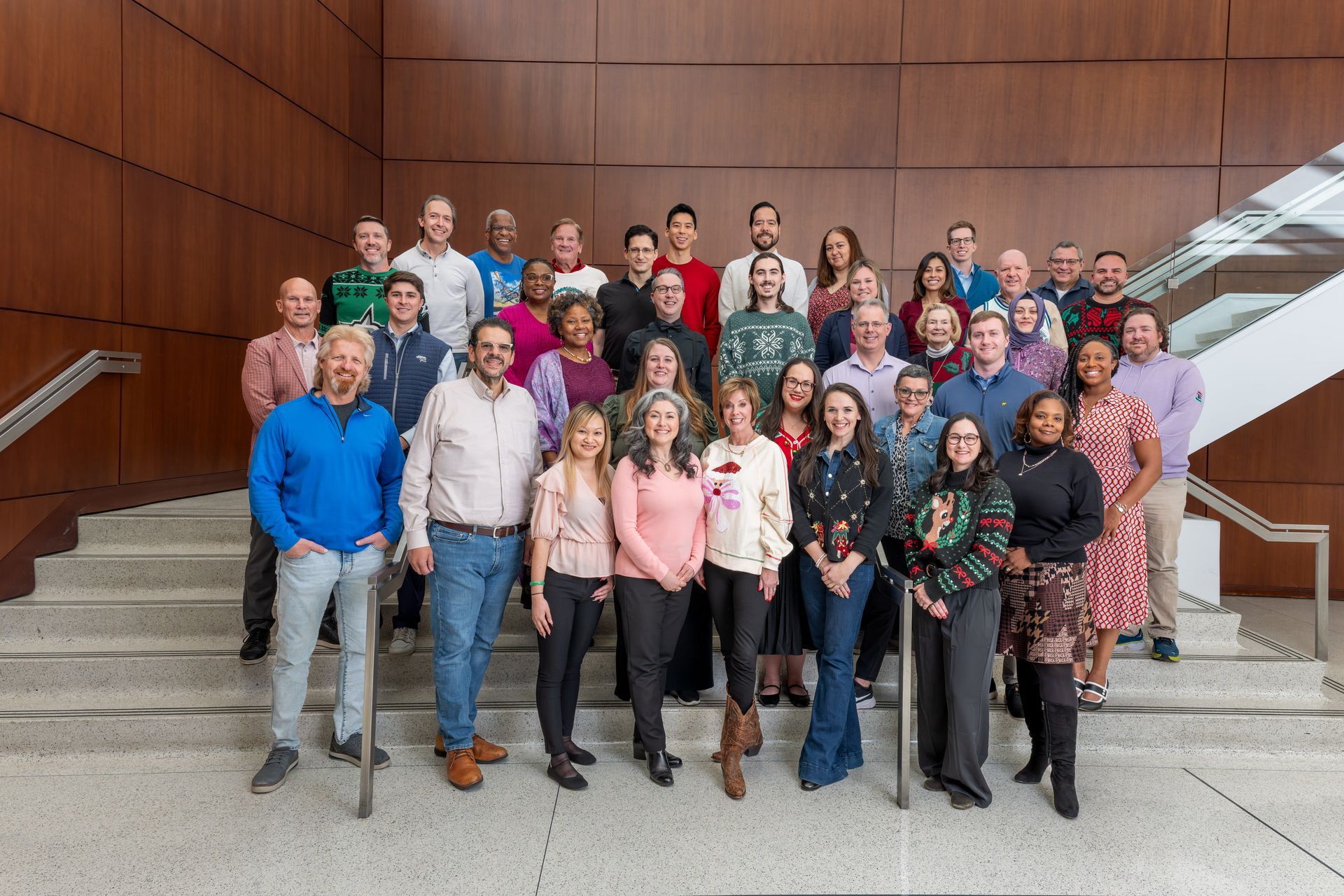 A large group of people stand together on indoor stairs in front of a wood-paneled wall, smiling for a professional photo.