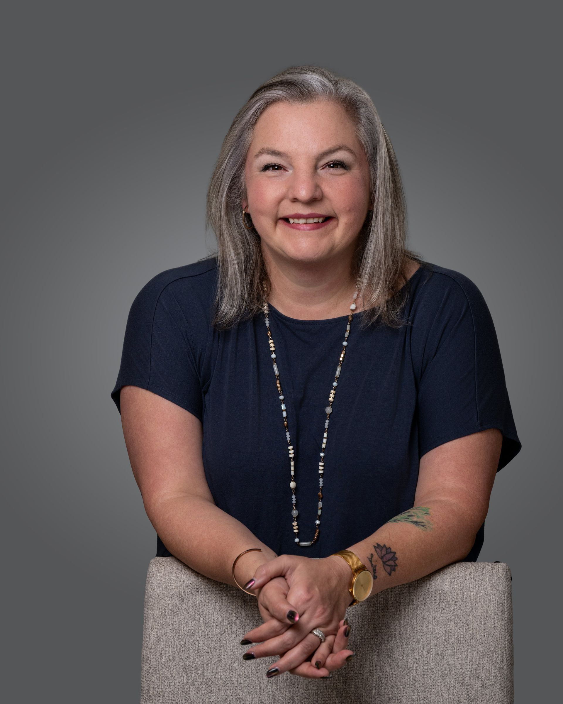 A smiling person wearing a dark blue shirt and beaded necklace leans their arms on the back of a textured chair.