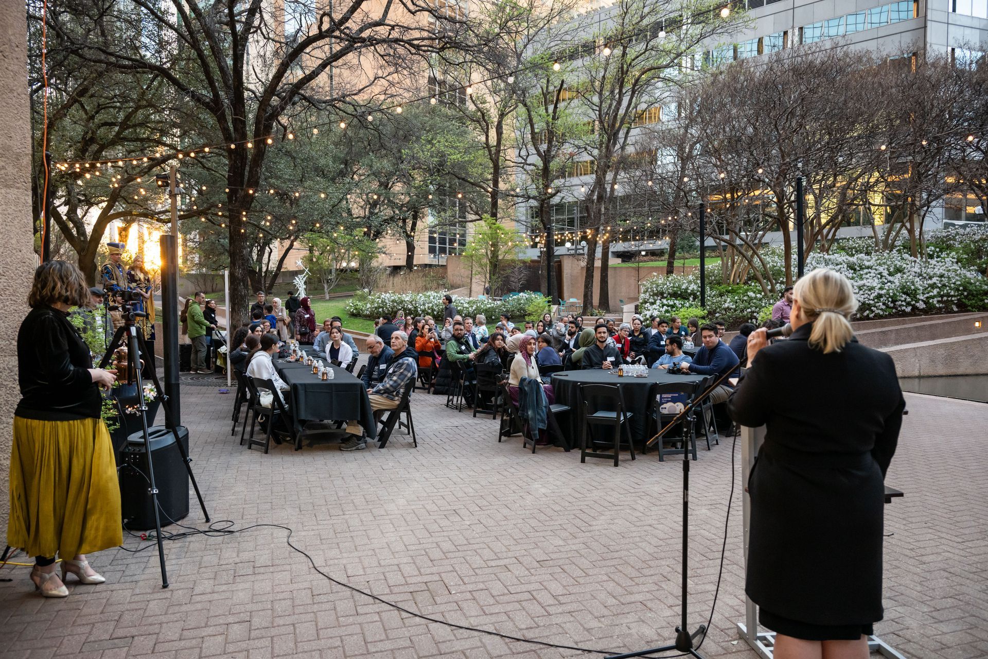 Speakers stand before a crowd seated at outdoor tables during an evening event in a tree-lined courtyard.