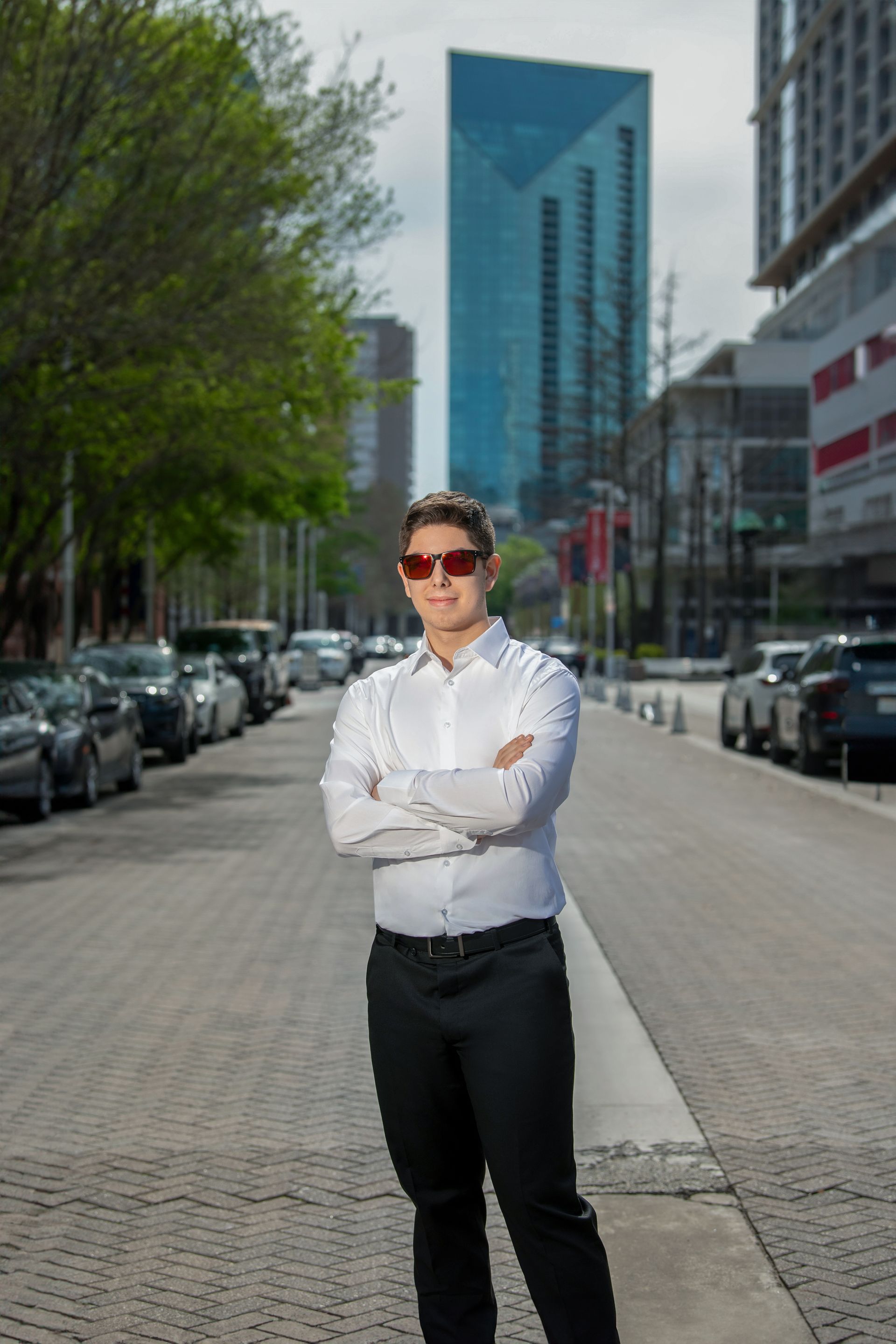 A graduate in a red cap and gown with a blue stole poses outside an auditorium entrance.