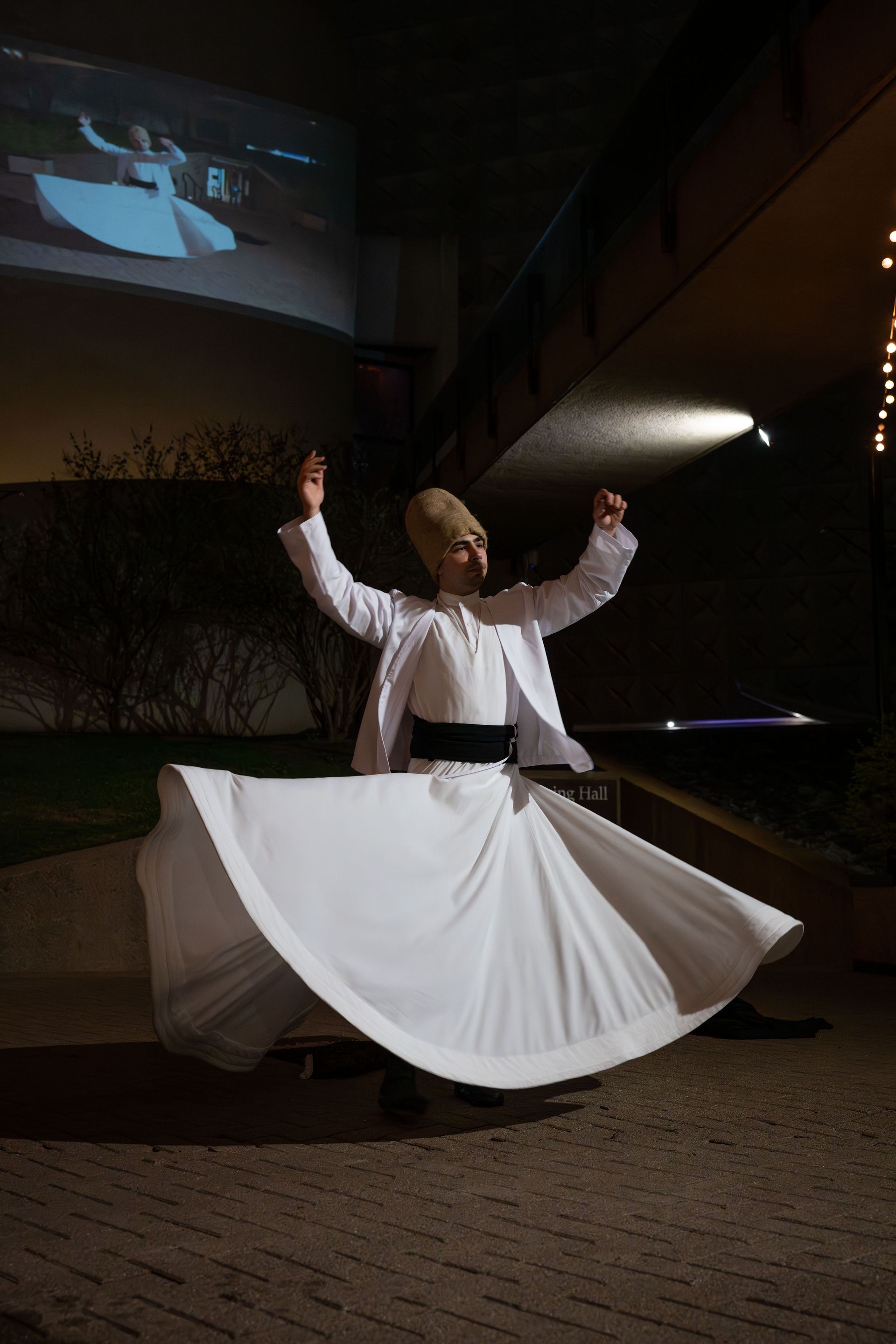 A whirling dervish in traditional white robes and a tall tan hat performs a meditative dance in a dimly lit setting.