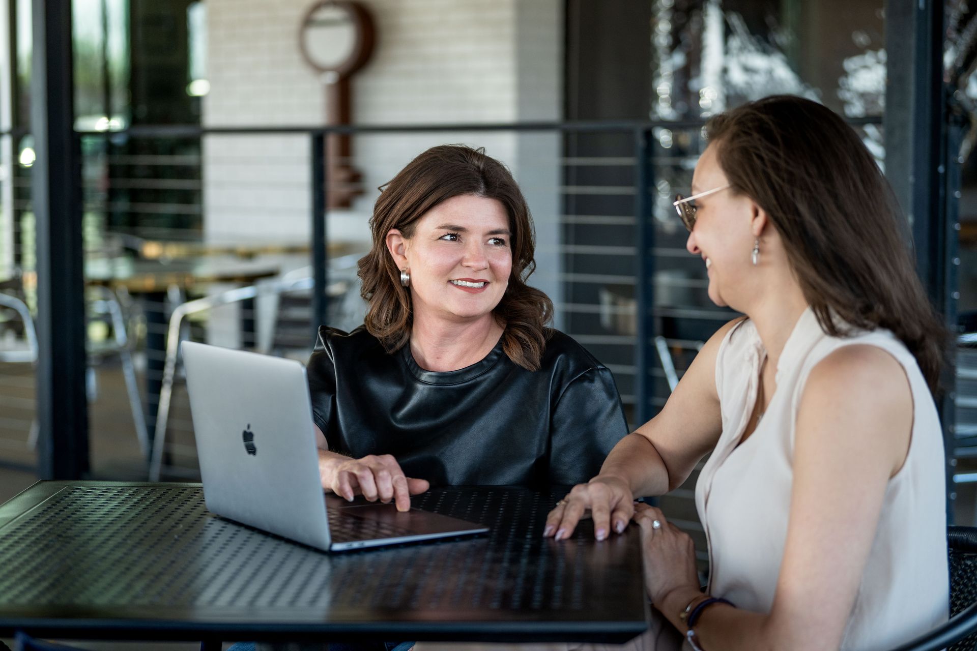 Two people sitting at an outdoor metal table with a laptop, engaged in conversation while looking at the screen.