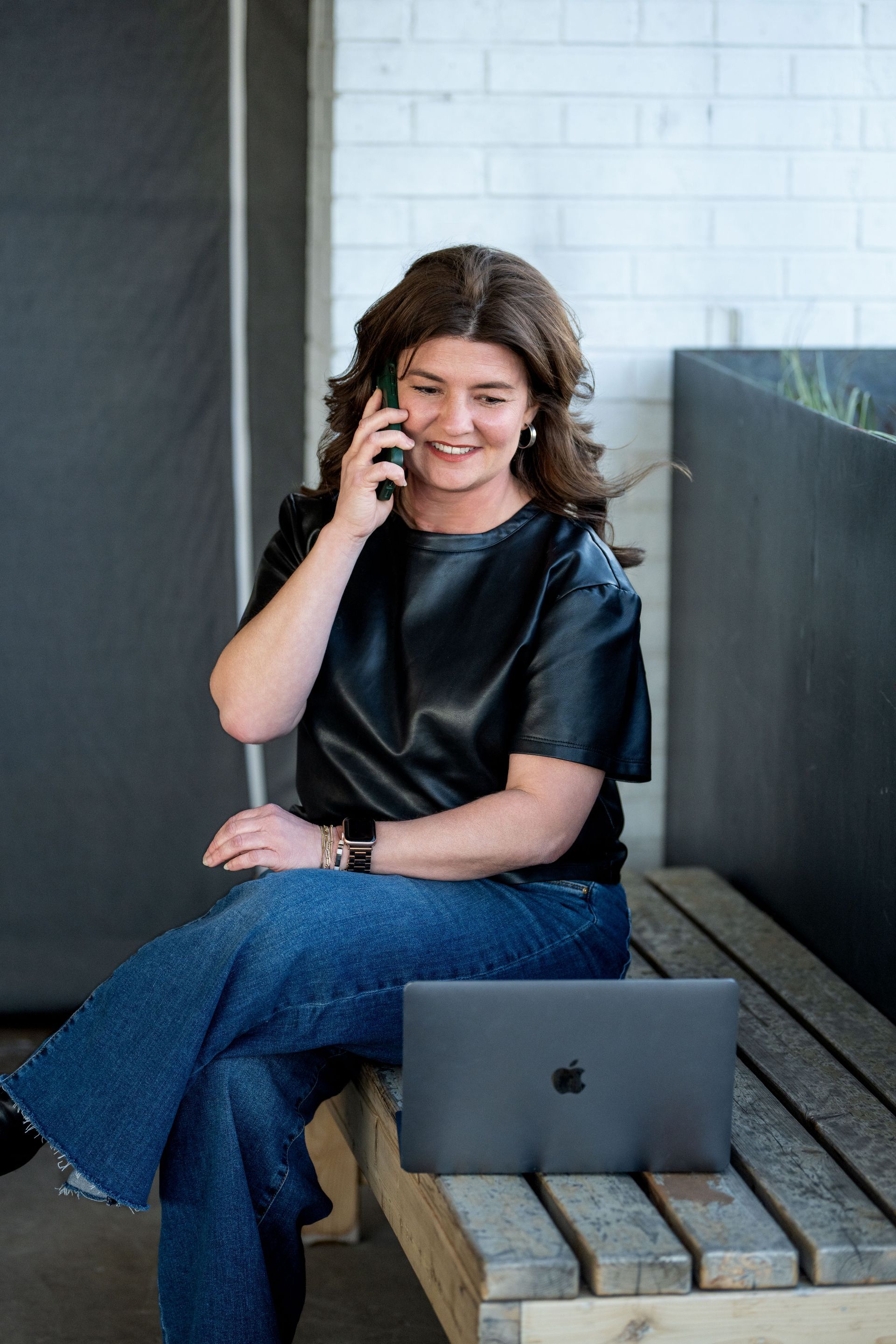 A person with wavy brown hair wearing a black shirt and jeans sits on a wooden bench, talking on a phone near a laptop.