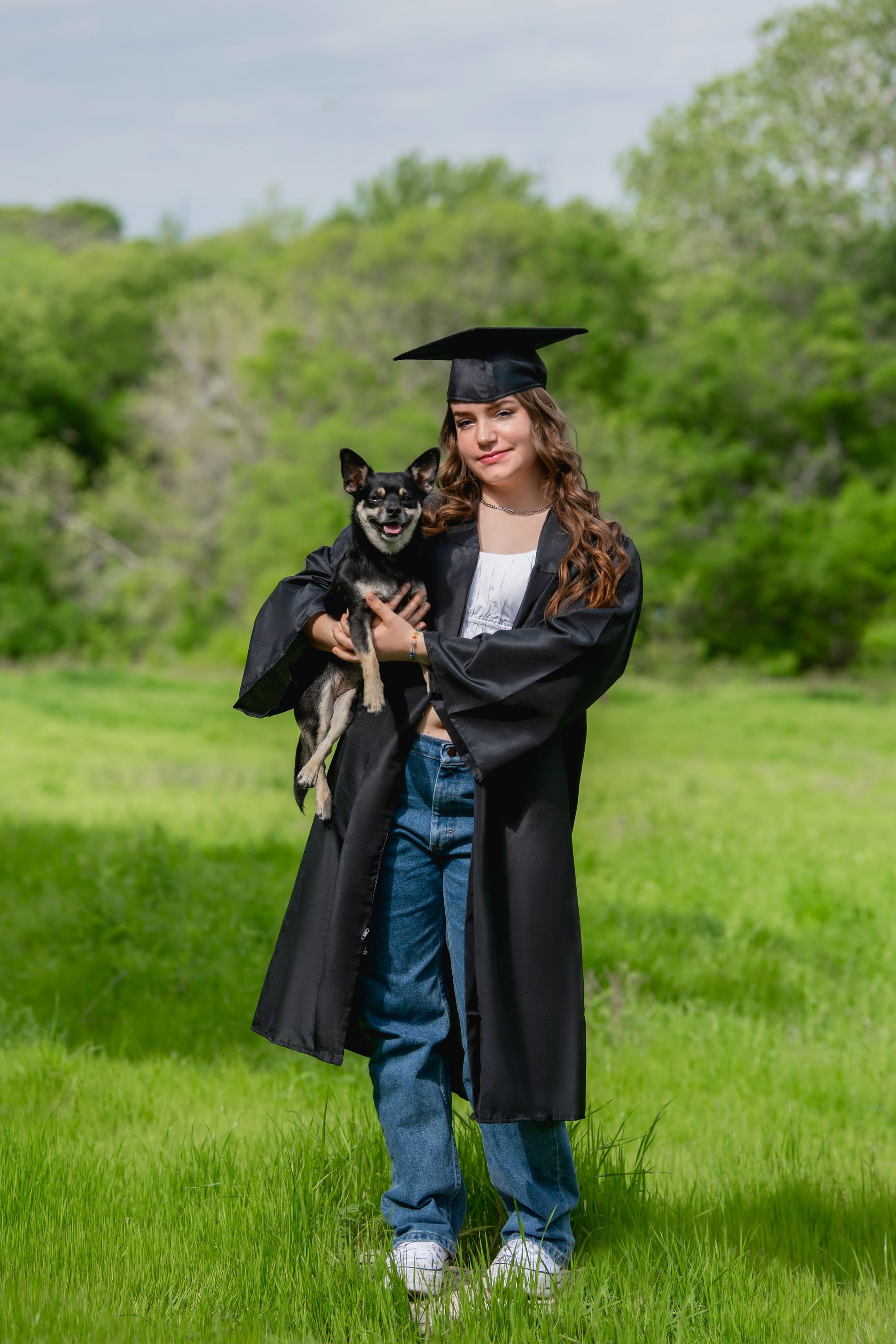 A person in a purple graduation gown and white stole stands outside under a pergola, holding a mortarboard.