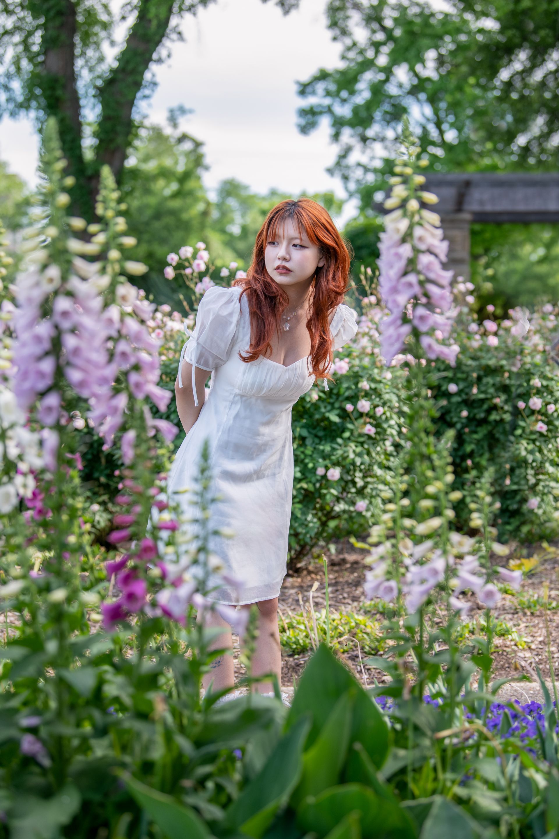 A smiling person kneeling by a lake, draped in a dark green plaid blanket, wearing jeans and tan wedge shoes.