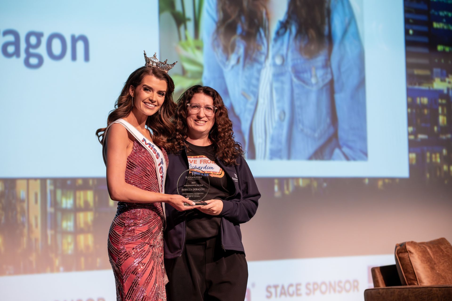 A woman wearing a pageant sash and crown stands onstage holding an award with another woman against a large backdrop.