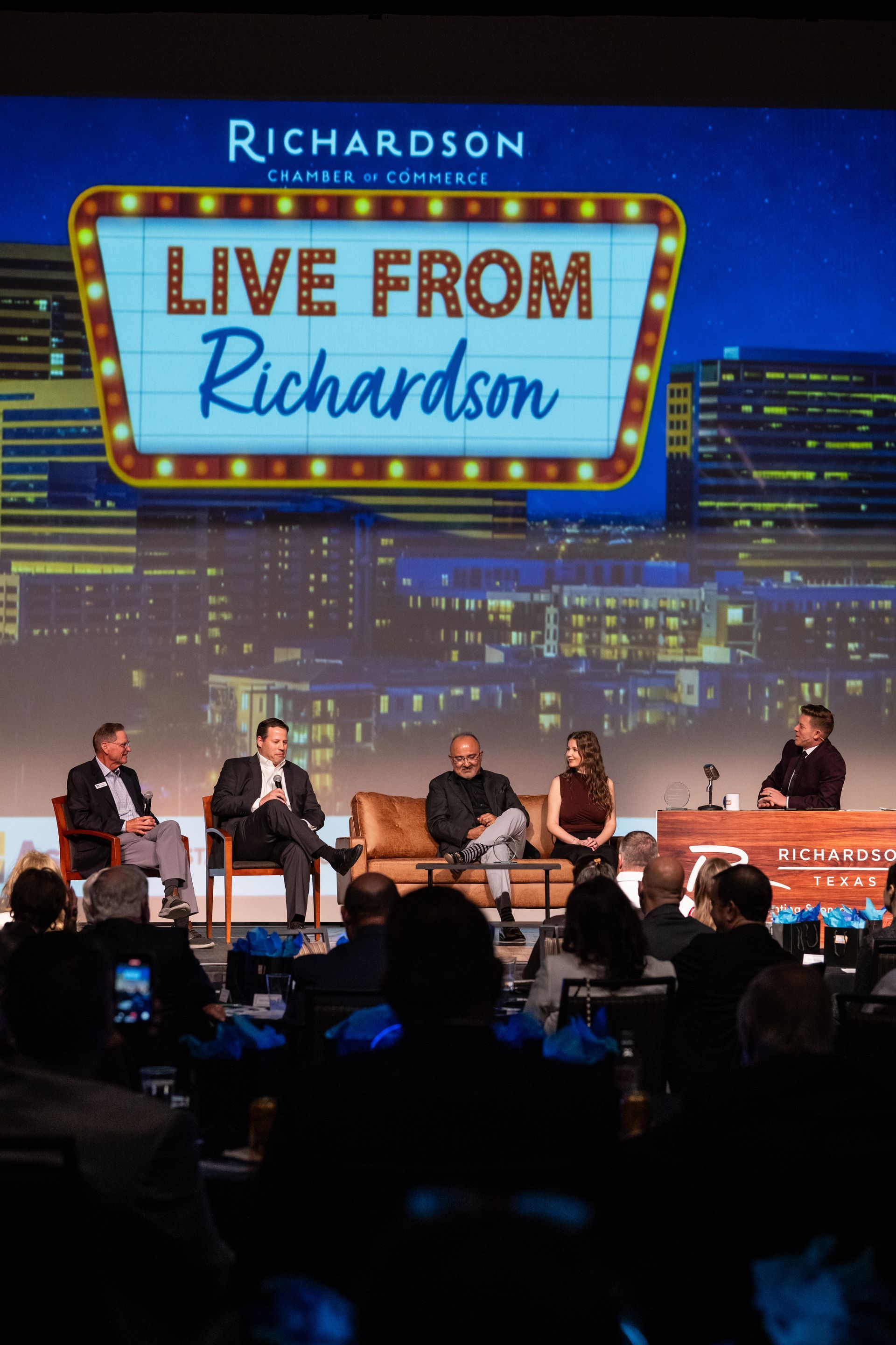 A panel of five people seated on a stage beneath a neon-style 