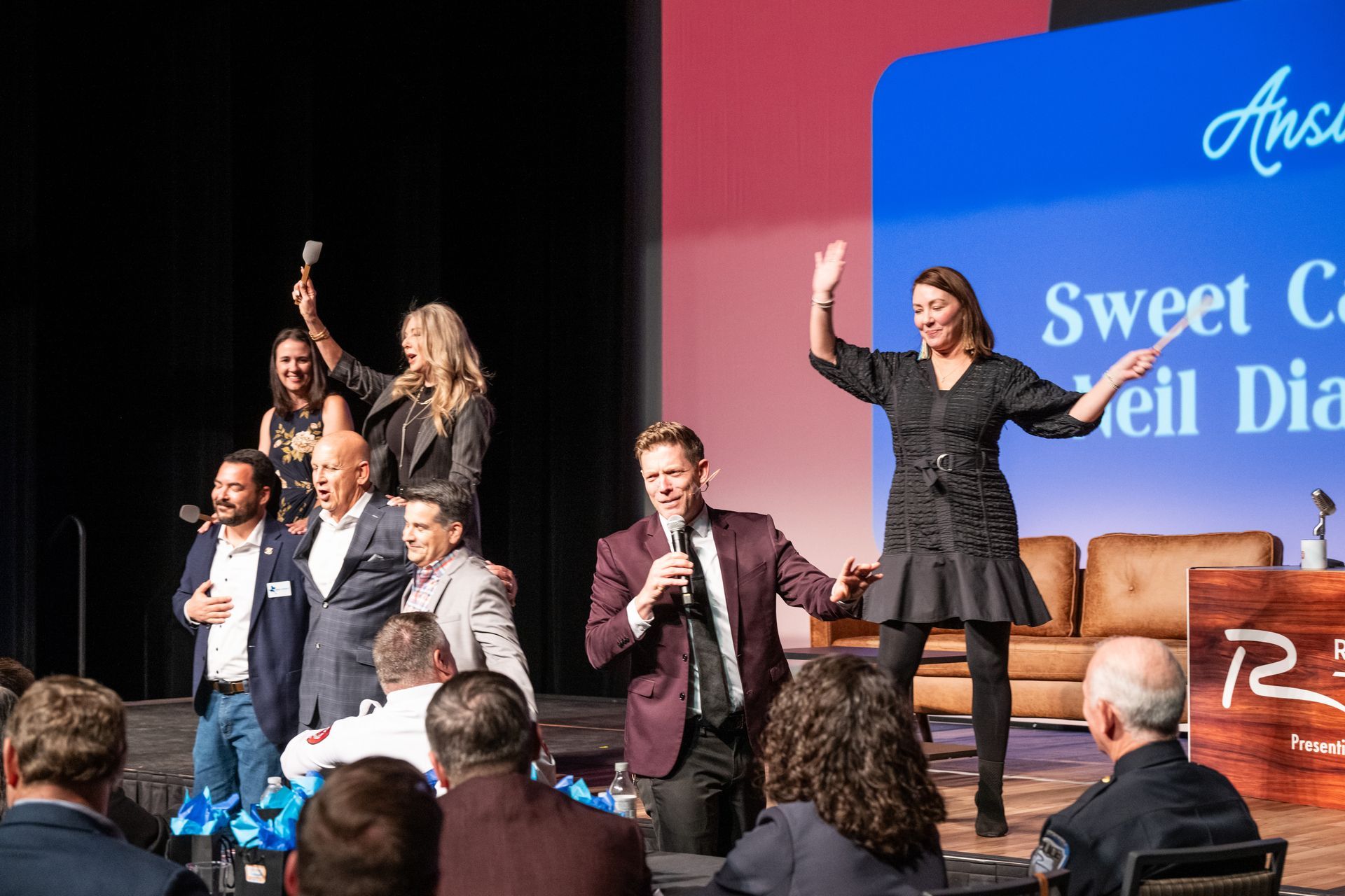 A group of people on a stage during an event, some waving and smiling, with a presentation screen in the background.