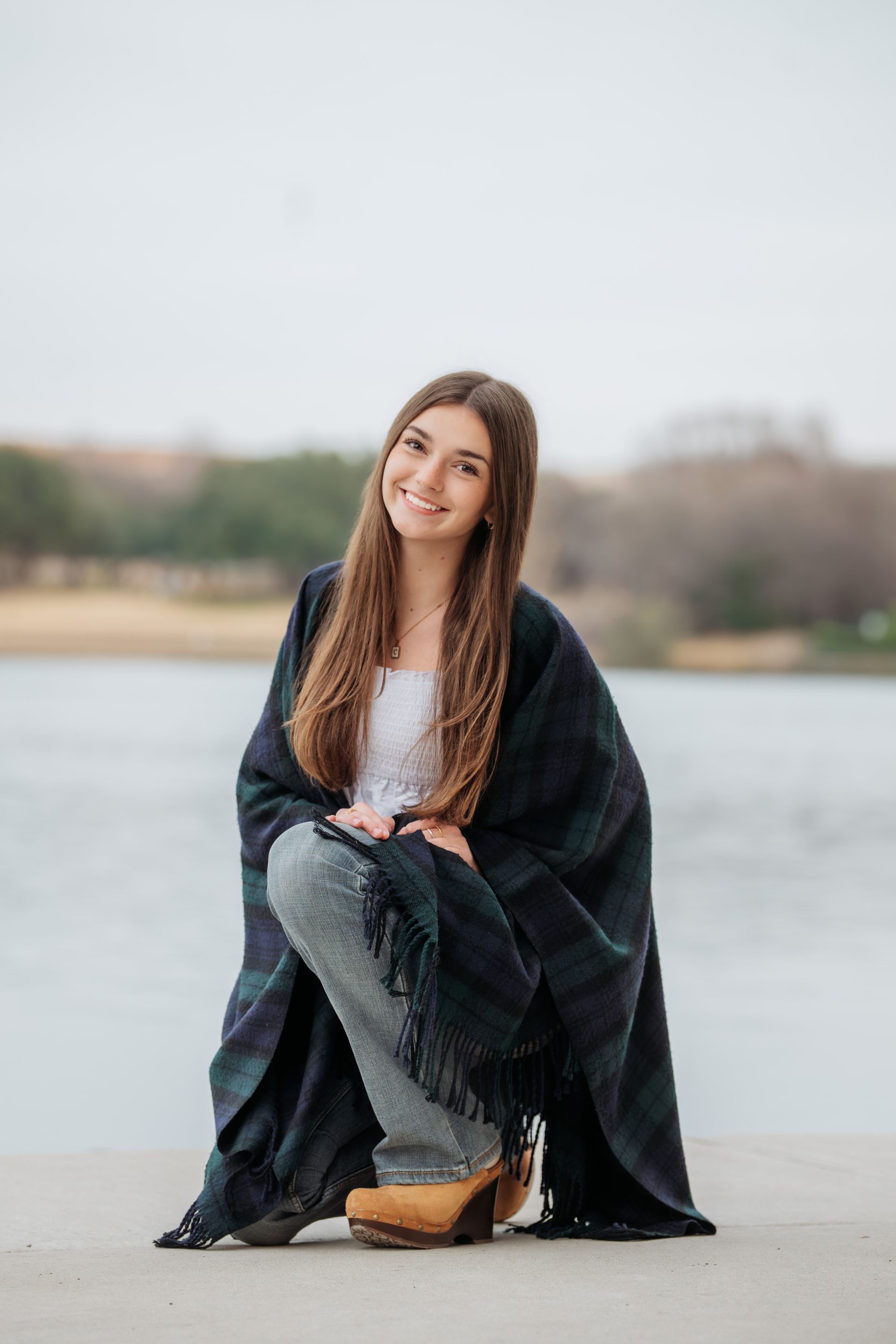 A smiling person wearing a dark plaid blanket and patterned pants, kneeling outdoors by a body of water.