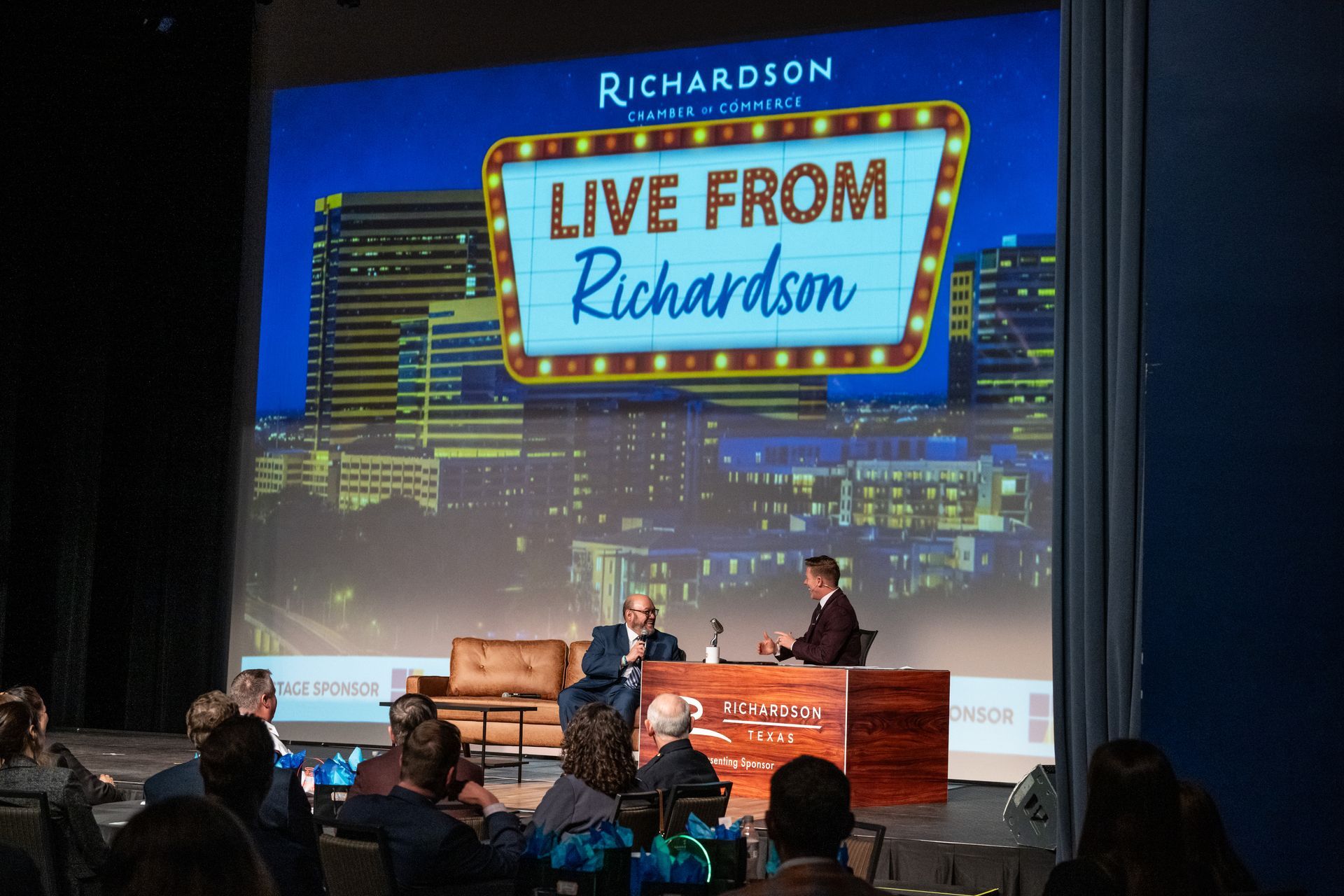 Two people sit on a stage during a presentation titled 