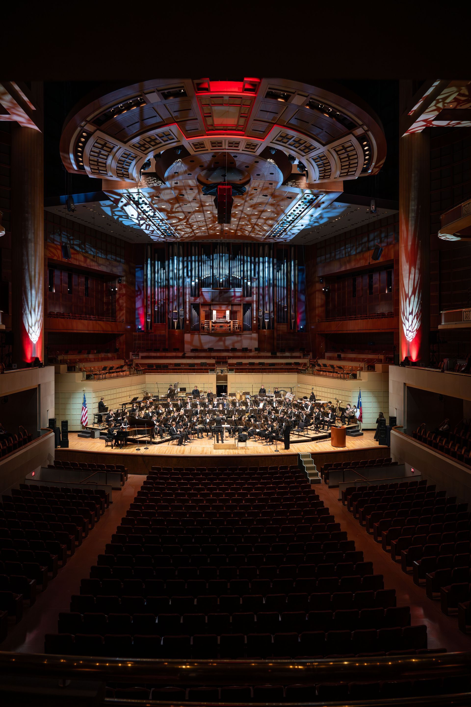 A large orchestra performs on a brightly lit stage in a grand concert hall with dark seating and an ornate ceiling.