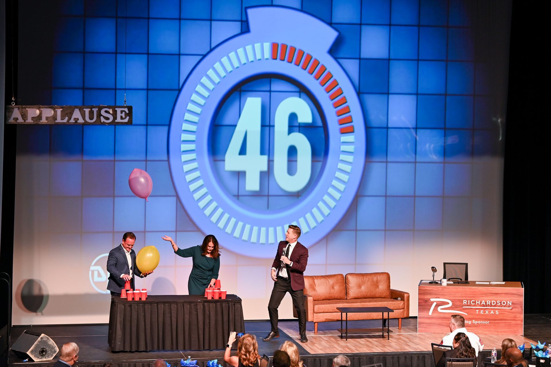 Three people compete in a balloon game on a stage in front of a large screen showing a countdown timer at 46.