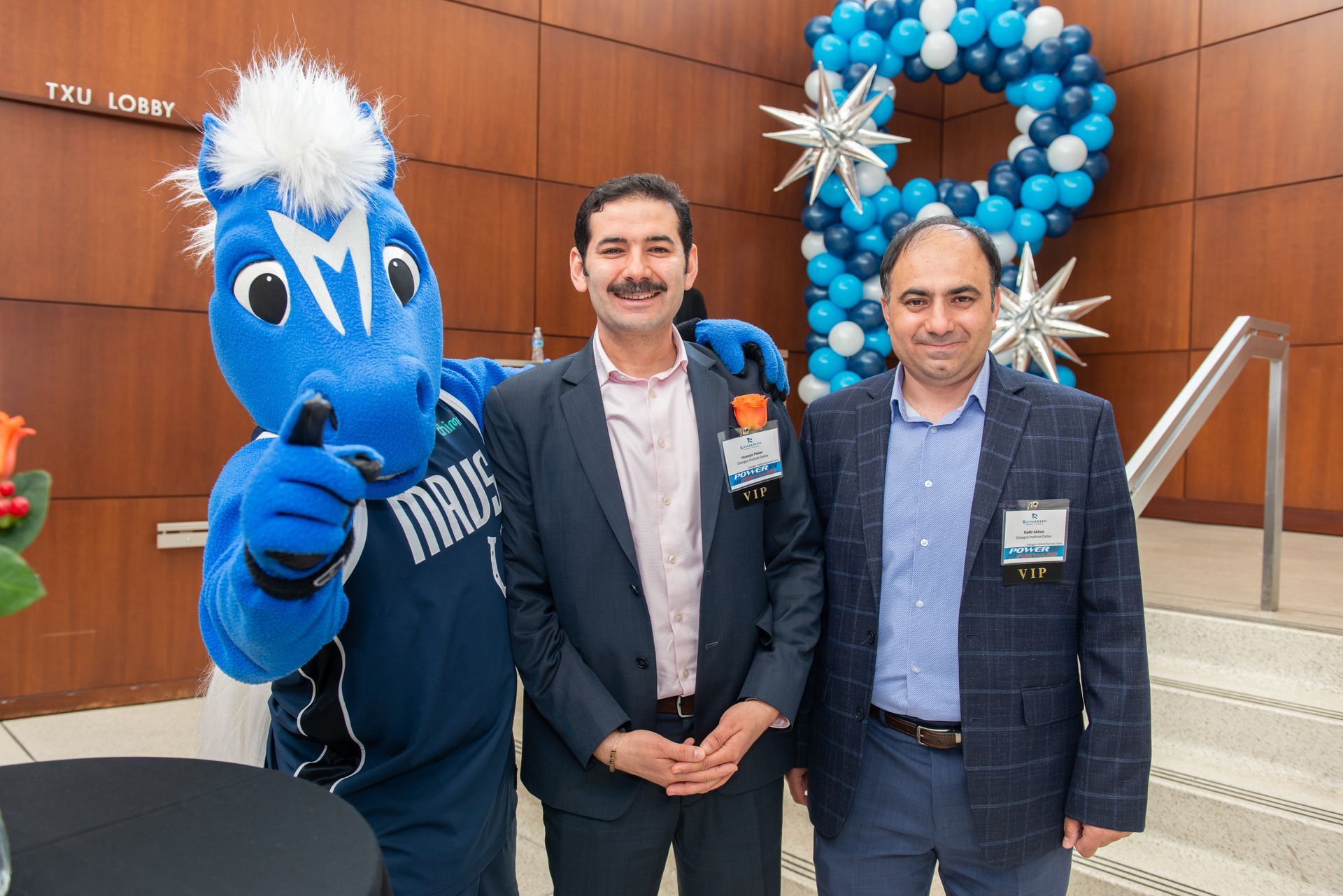 A blue horse mascot poses with two men in business attire in front of a blue balloon display in a lobby.