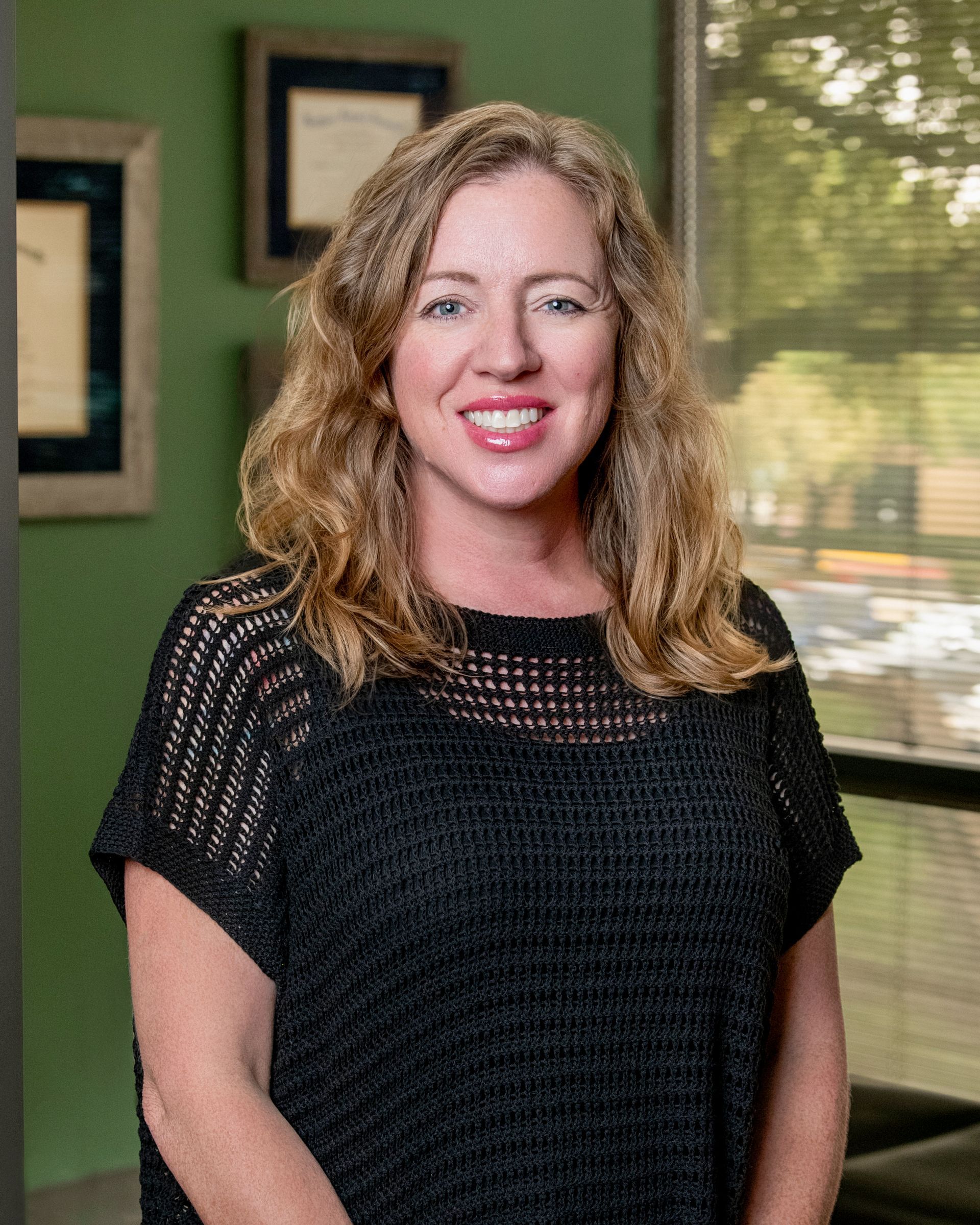 A person with shoulder-length wavy hair and a smile, wearing a black open-knit top in an office setting.