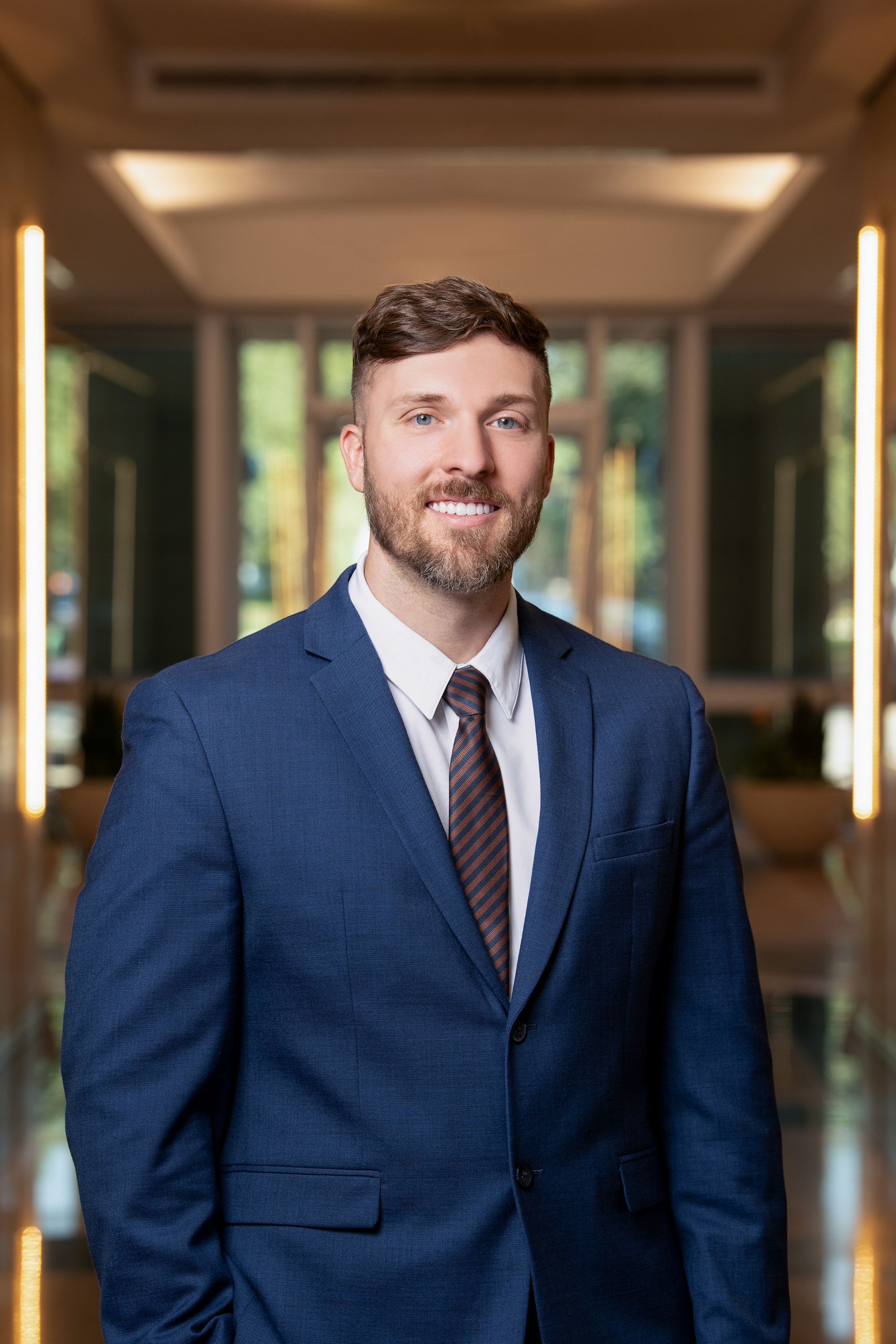 A professional portrait of a person in a blue blazer, white shirt, and patterned tie, standing in a brightly lit hallway.