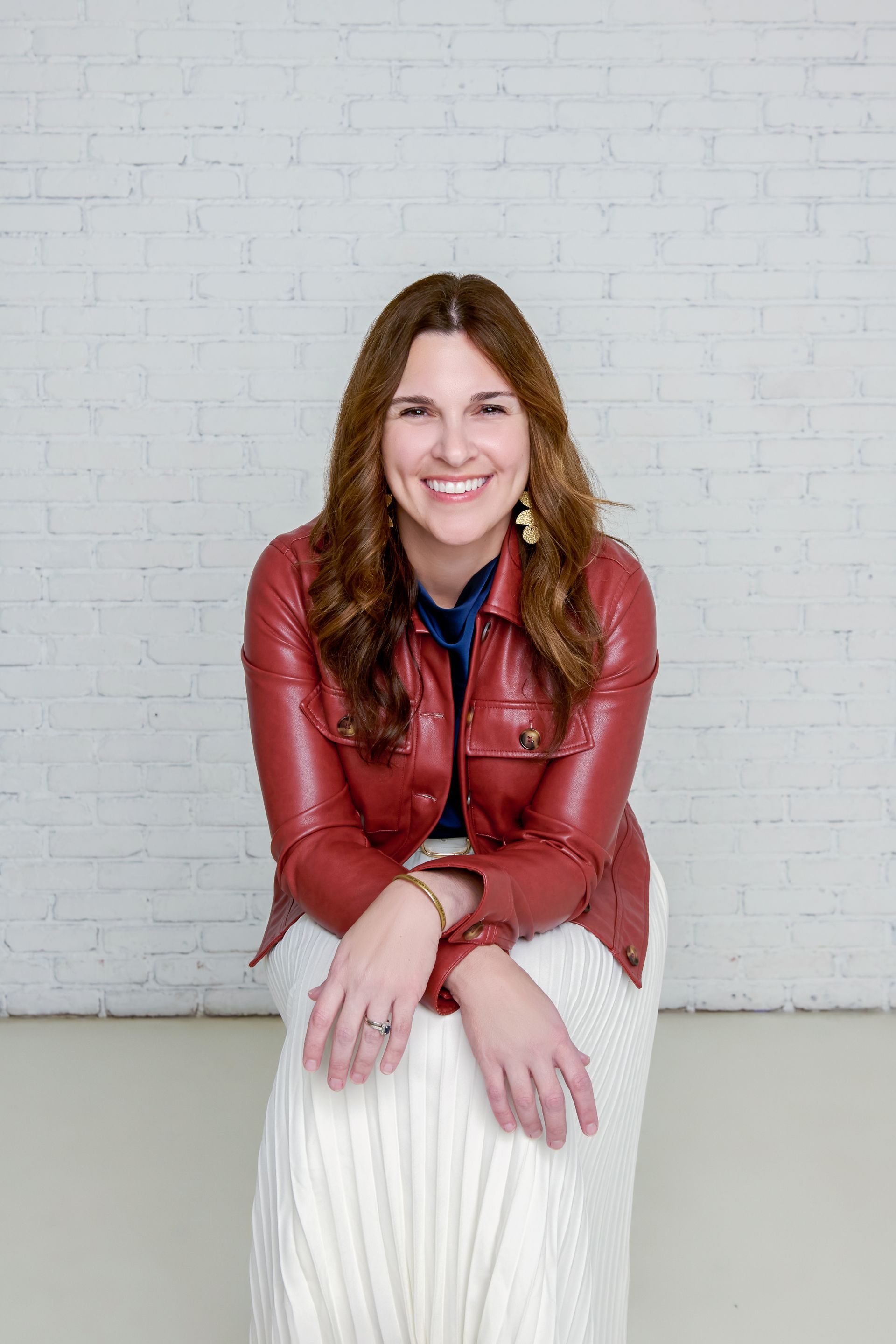 A smiling person wearing a red leather jacket and a long, cream-colored skirt sits against a white brick wall.
