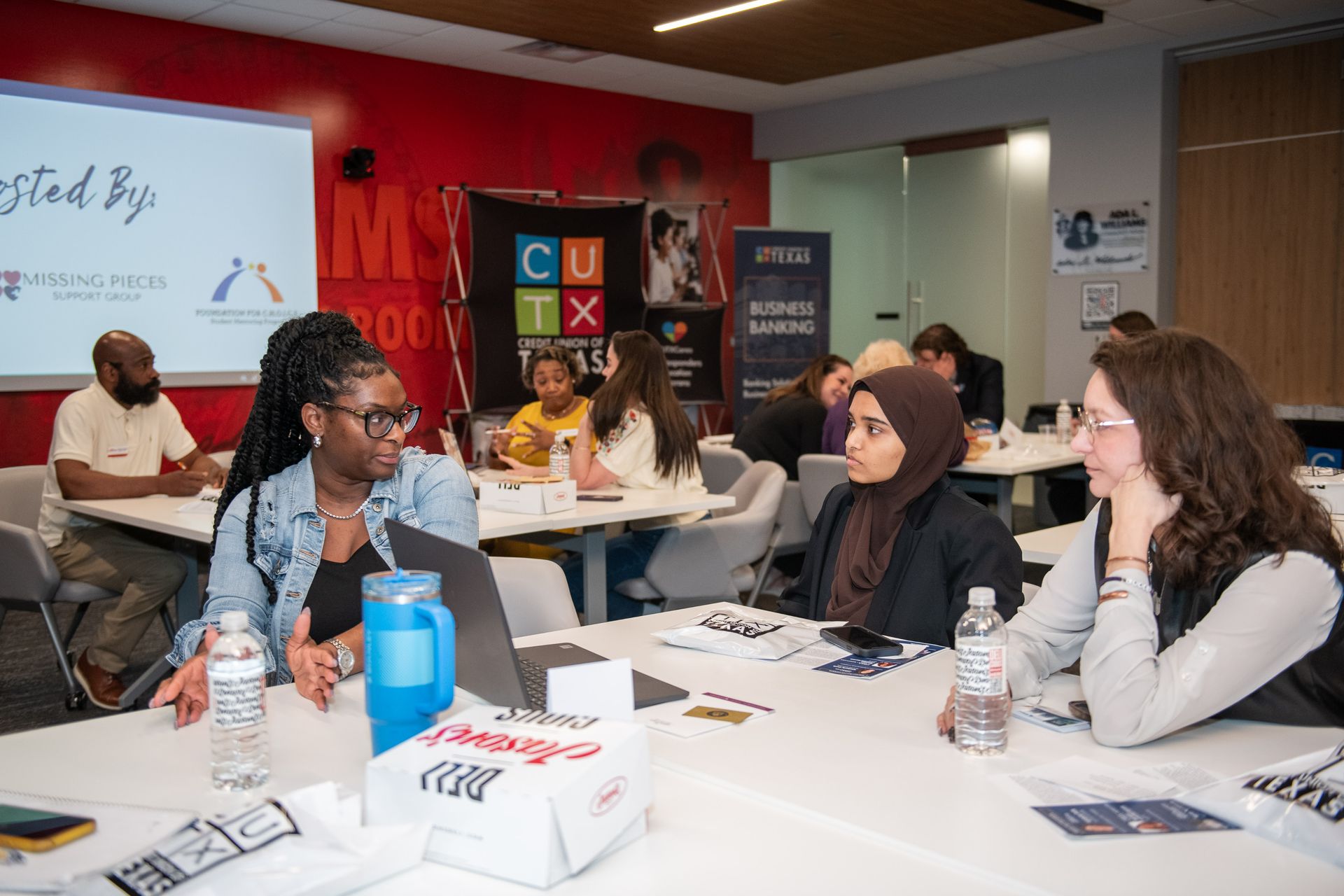 People sit at tables in a room with a red wall, participating in a business or workshop session with a digital display.