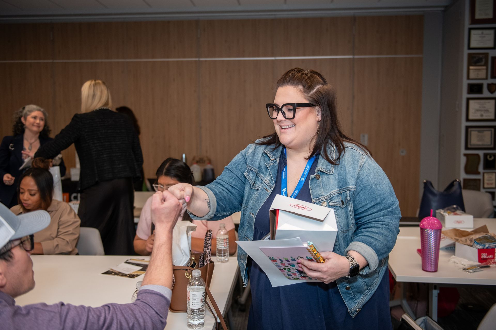 A woman wearing a denim jacket smiles while fist-bumping a person in a meeting room with people in the background.