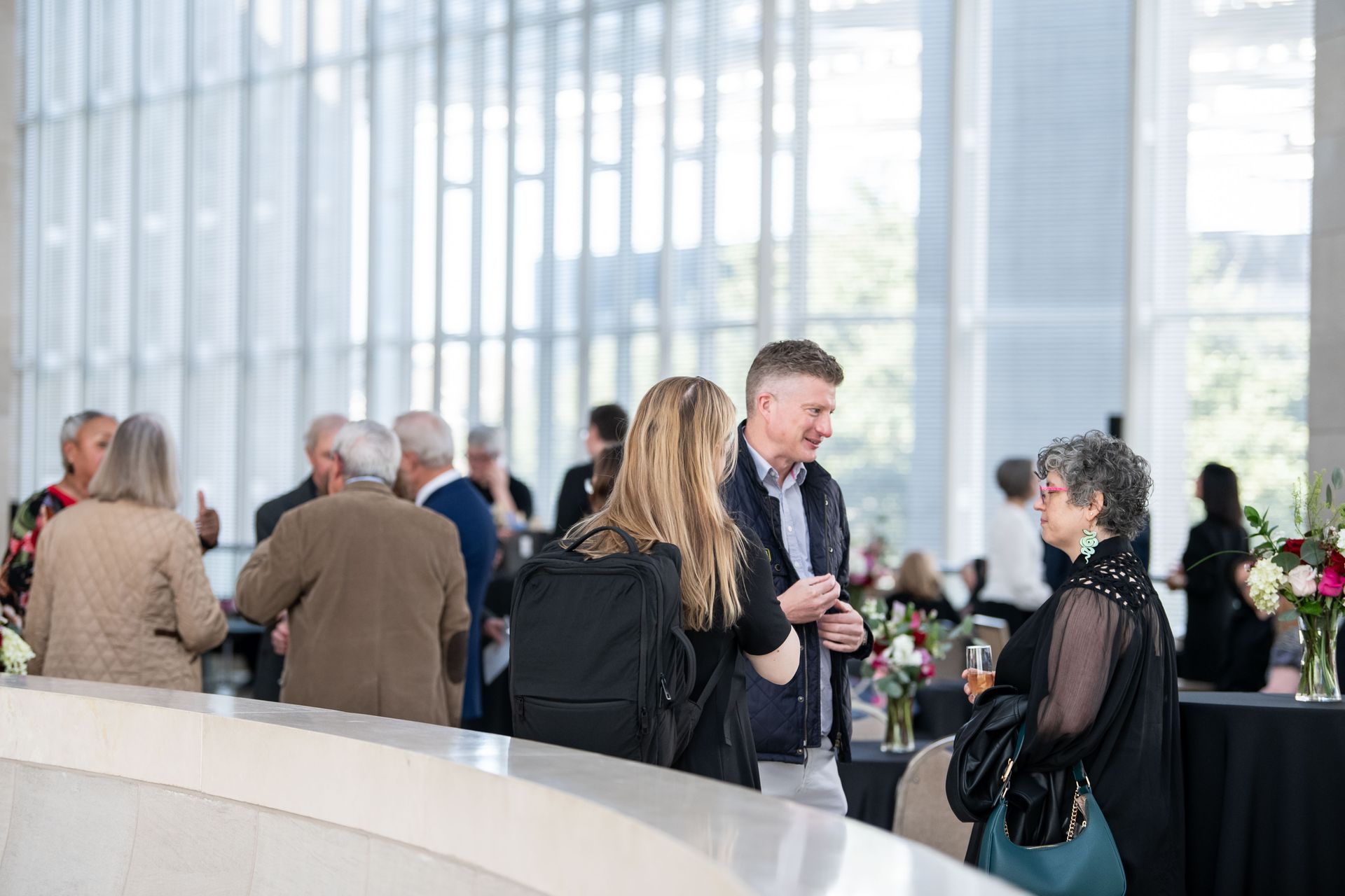 People in business-casual attire talk and socialize during an indoor event in a modern hall with floor-to-ceiling windows.
