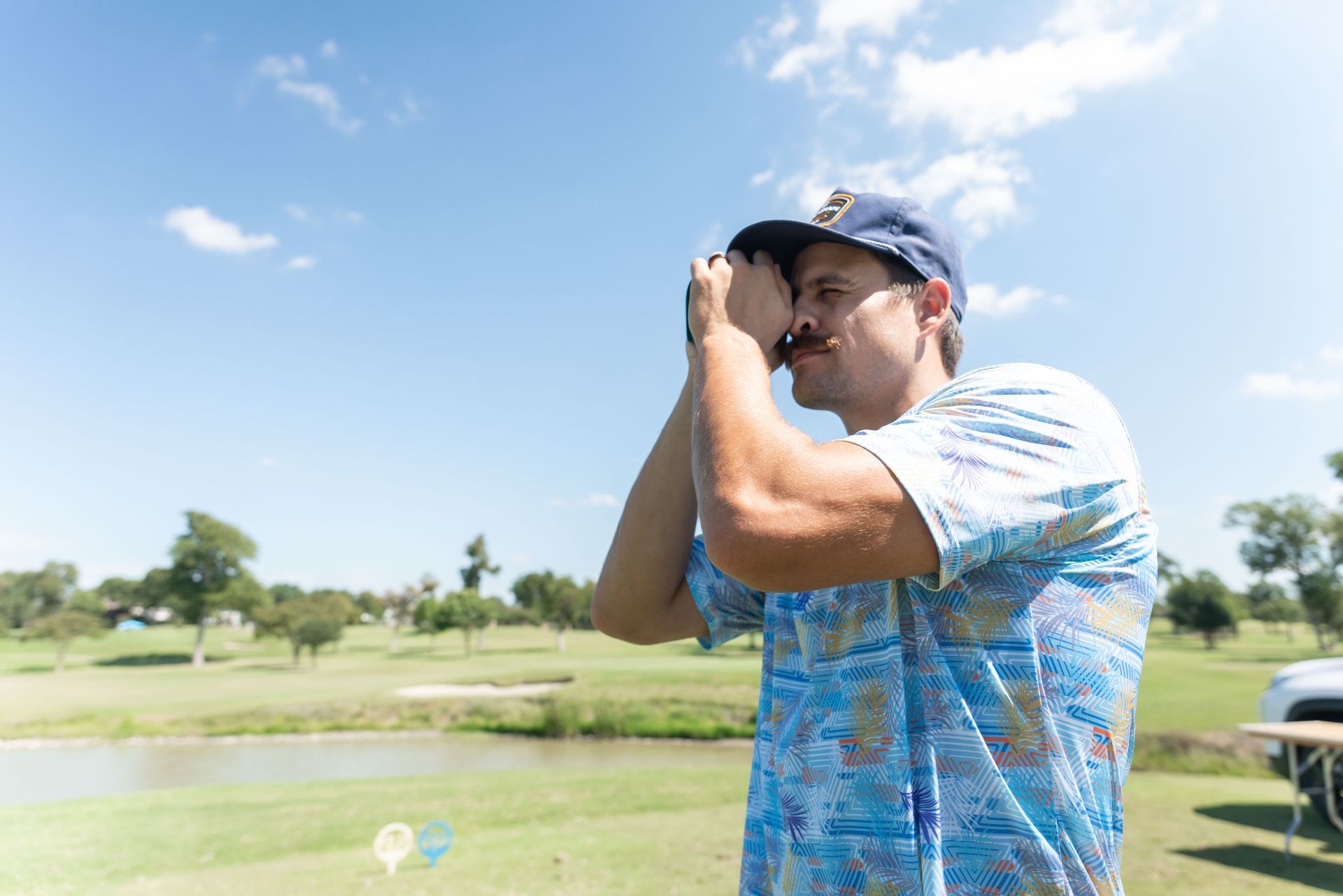 A person with a mustache, wearing a blue cap and patterned shirt, looking through binoculars on a sunny golf course.