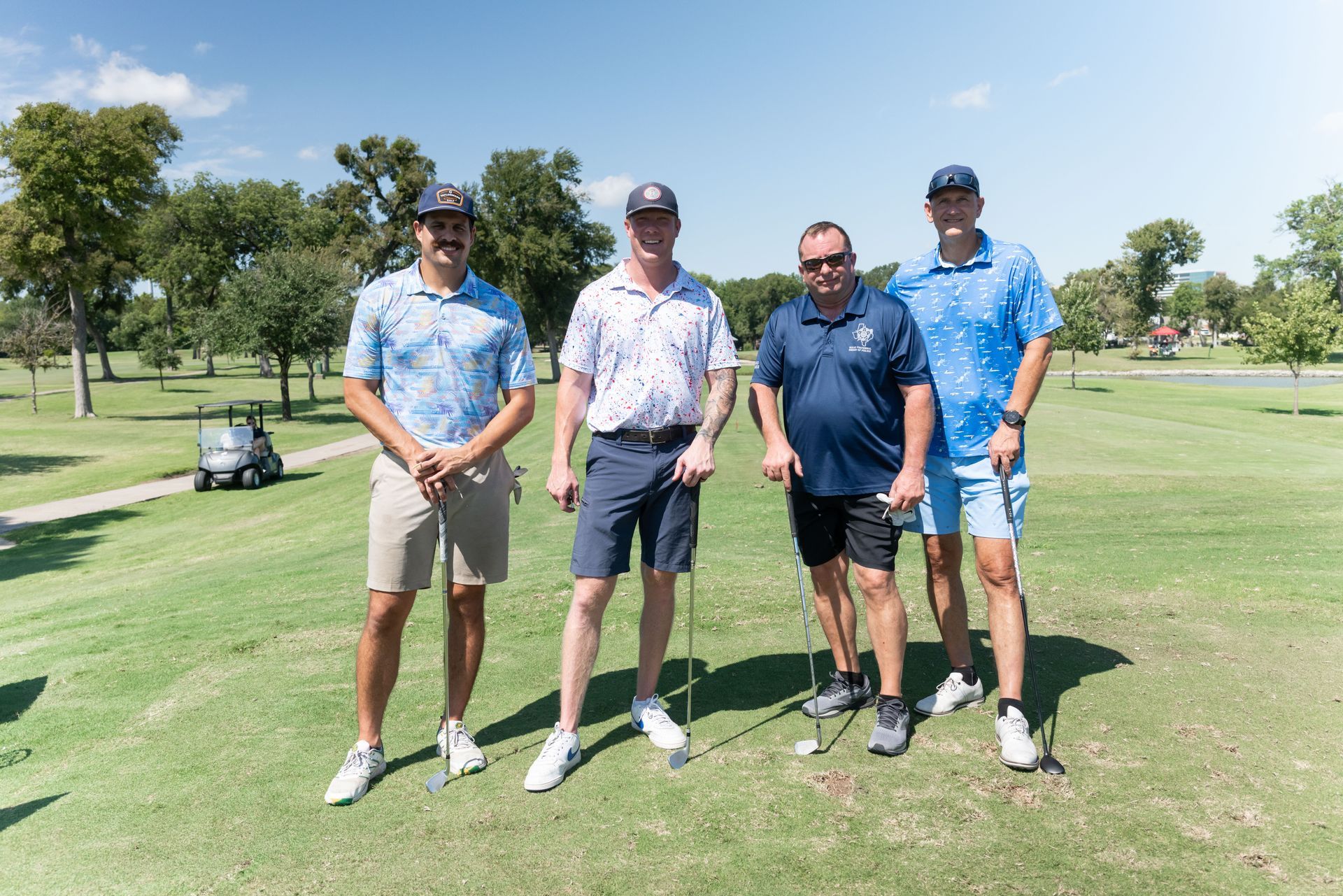 Four golfers in shirts and shorts stand on a sunny green course, posing together with golf clubs.