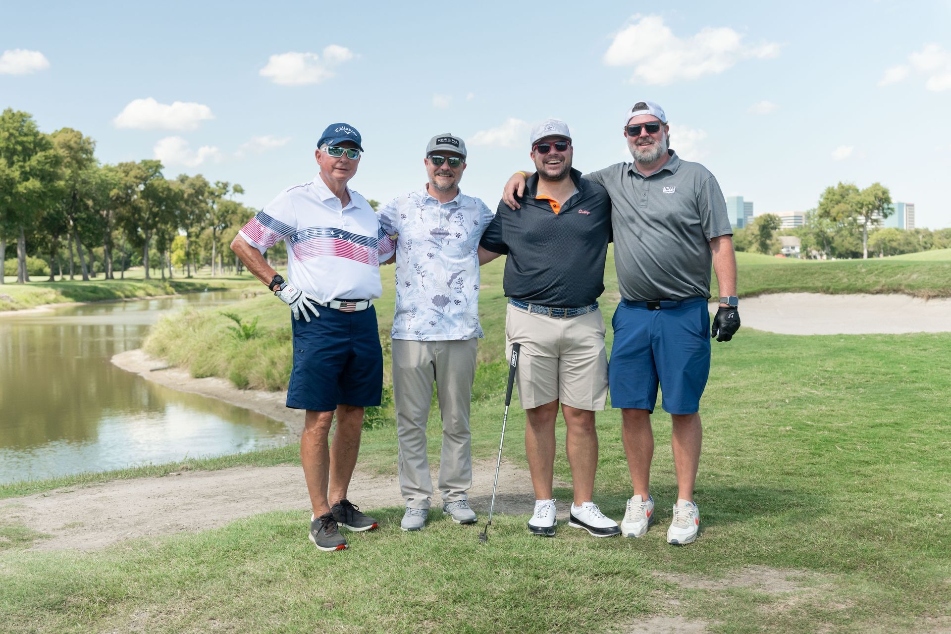 Four people stand together on a grassy golf course next to a water hazard, smiling for a group photo.