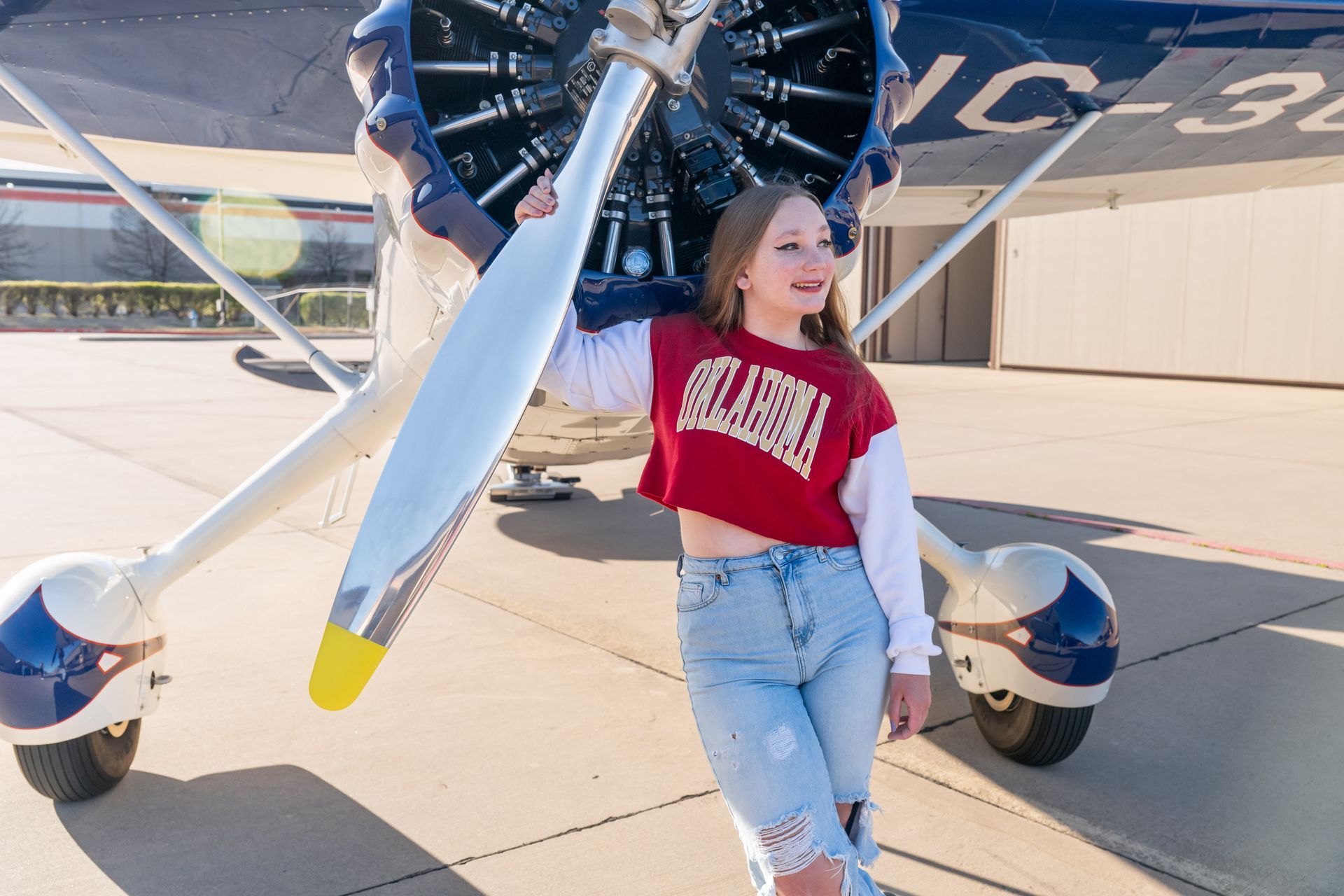 A person leaning against the propeller of a vintage aircraft outdoors, wearing a red University of Oklahoma sweatshirt.