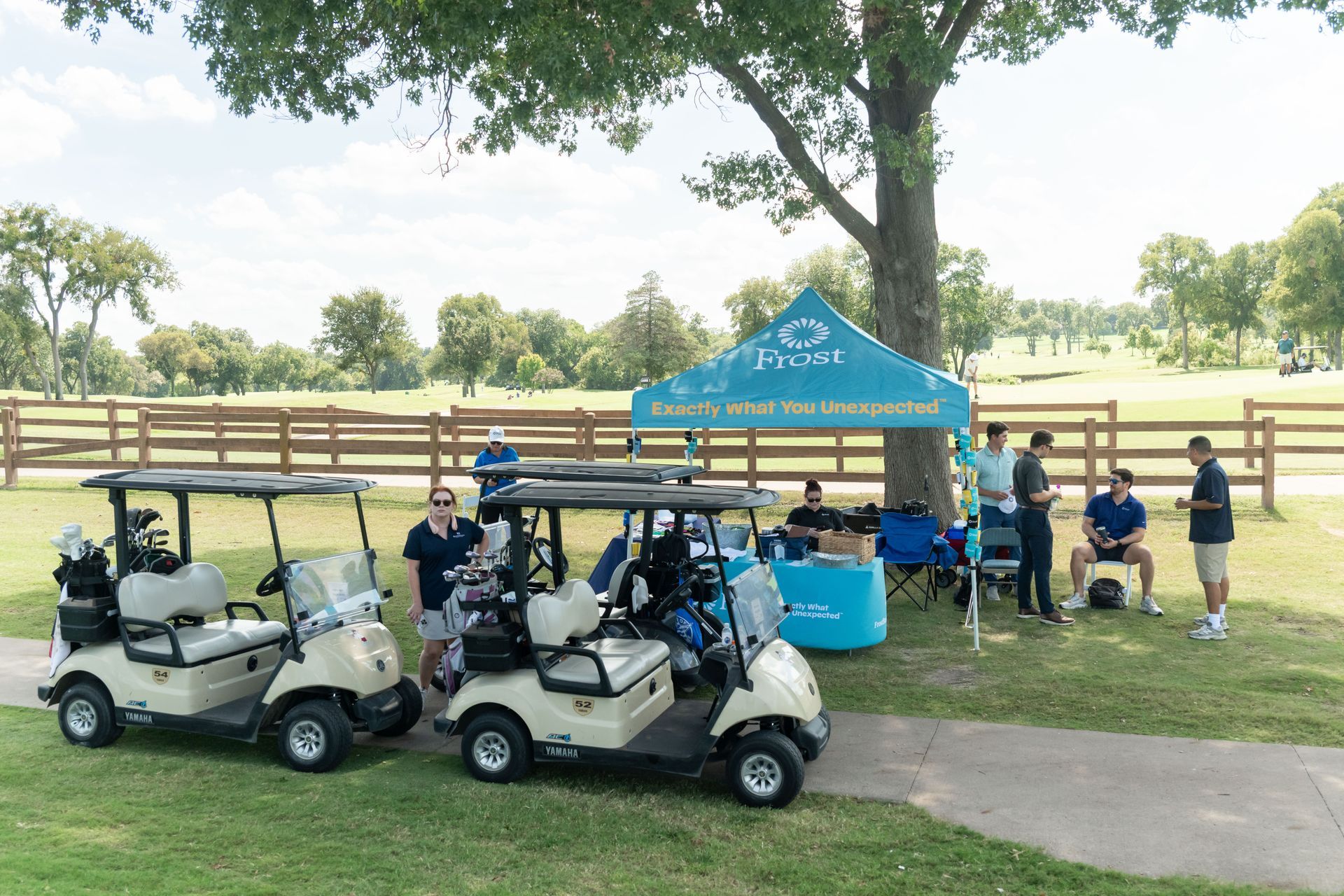 Two golf carts are parked on grass in front of a blue tent on a golf course with people gathered under the canopy.