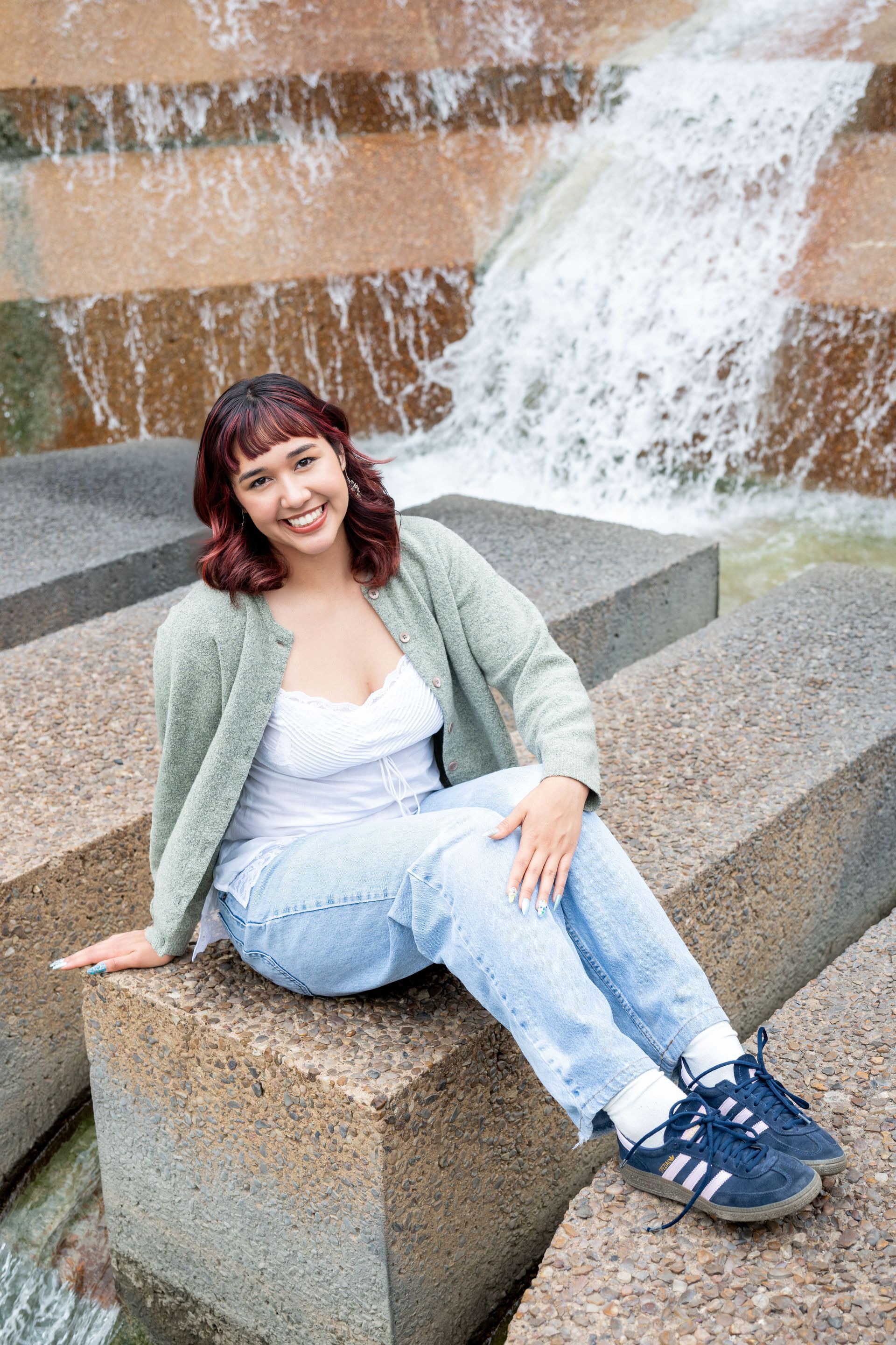 A smiling person sits on a stone block in front of a waterfall, wearing a green cardigan, white top, and jeans.
