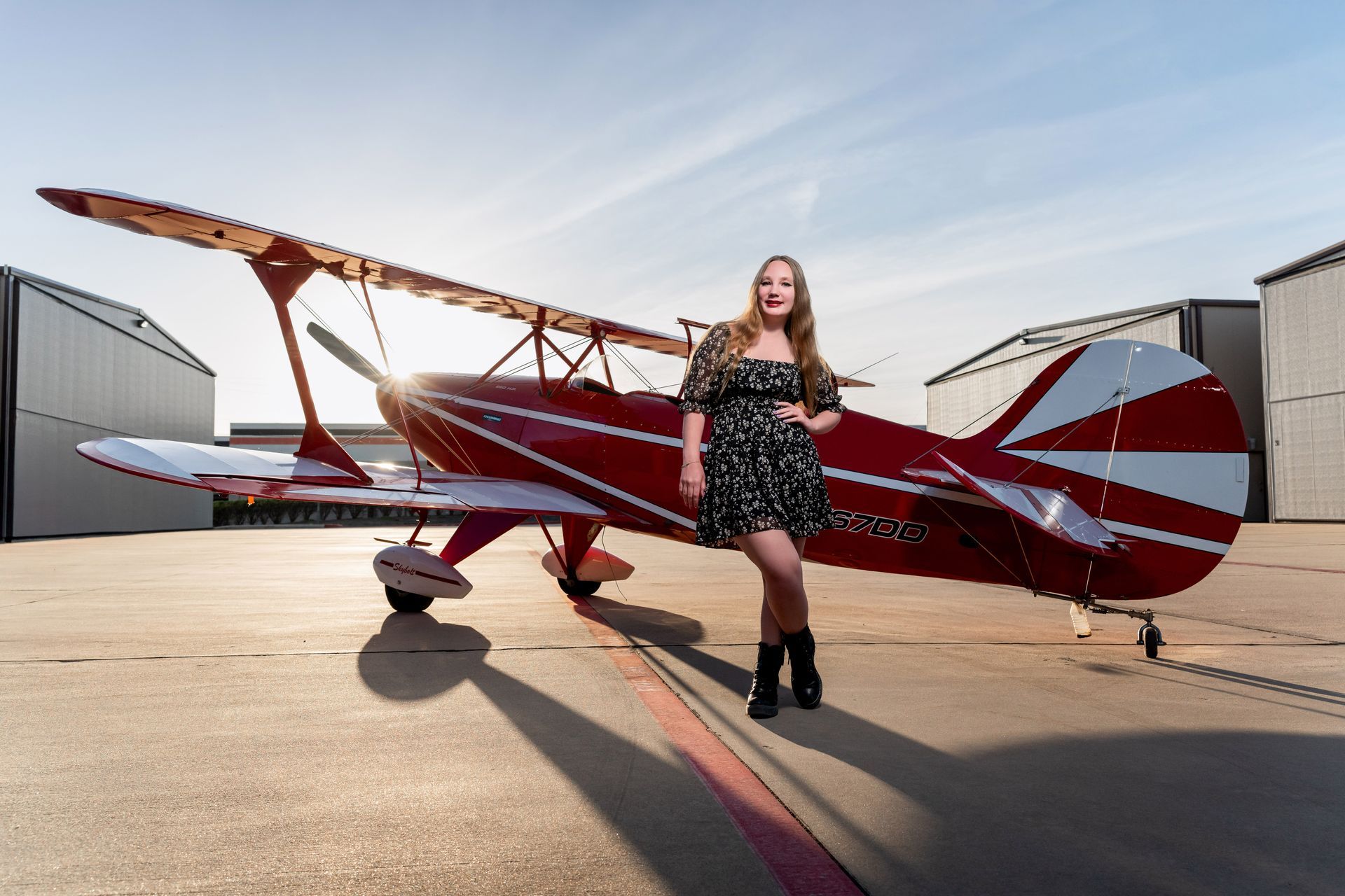 A woman in a floral dress stands posing in front of a red and white biplane on an airport tarmac near hangars.