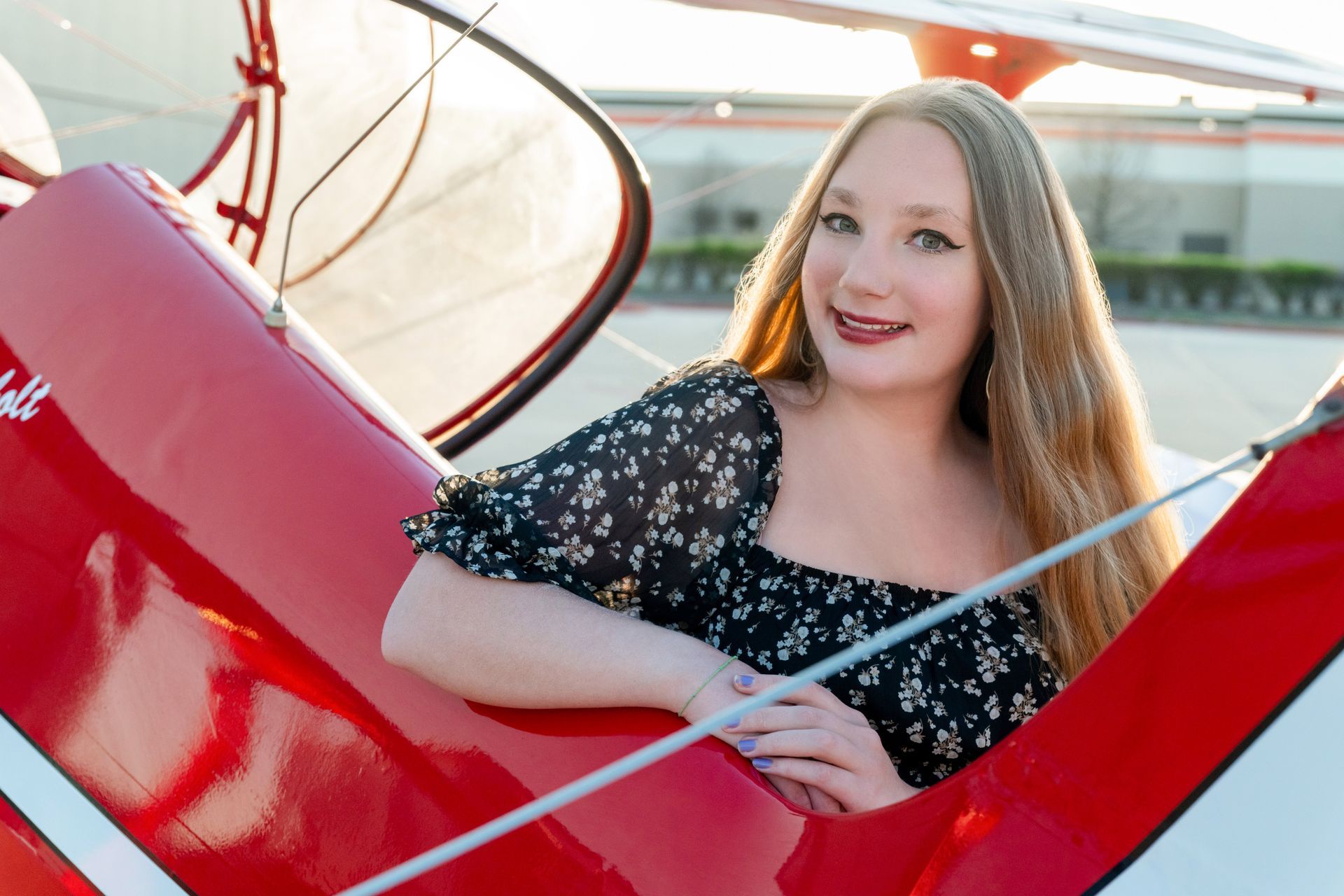 A person with long hair smiles while leaning into the cockpit of a bright red vintage airplane on a sunny day.