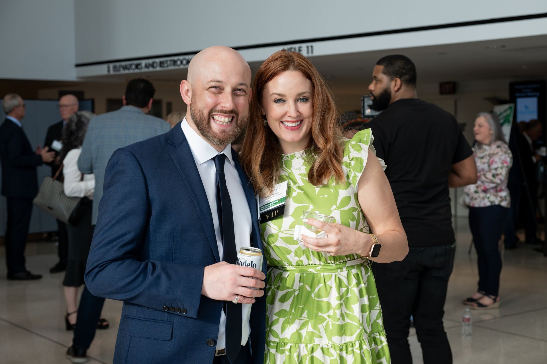 A smiling man in a suit and a woman in a green floral dress pose for a photo at a professional indoor event.